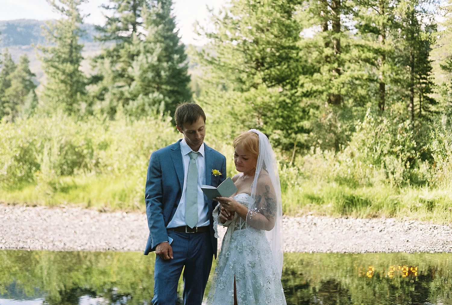 Bride and groom exchange vows outdoors by a serene pond surrounded by lush green trees on a sunny day.