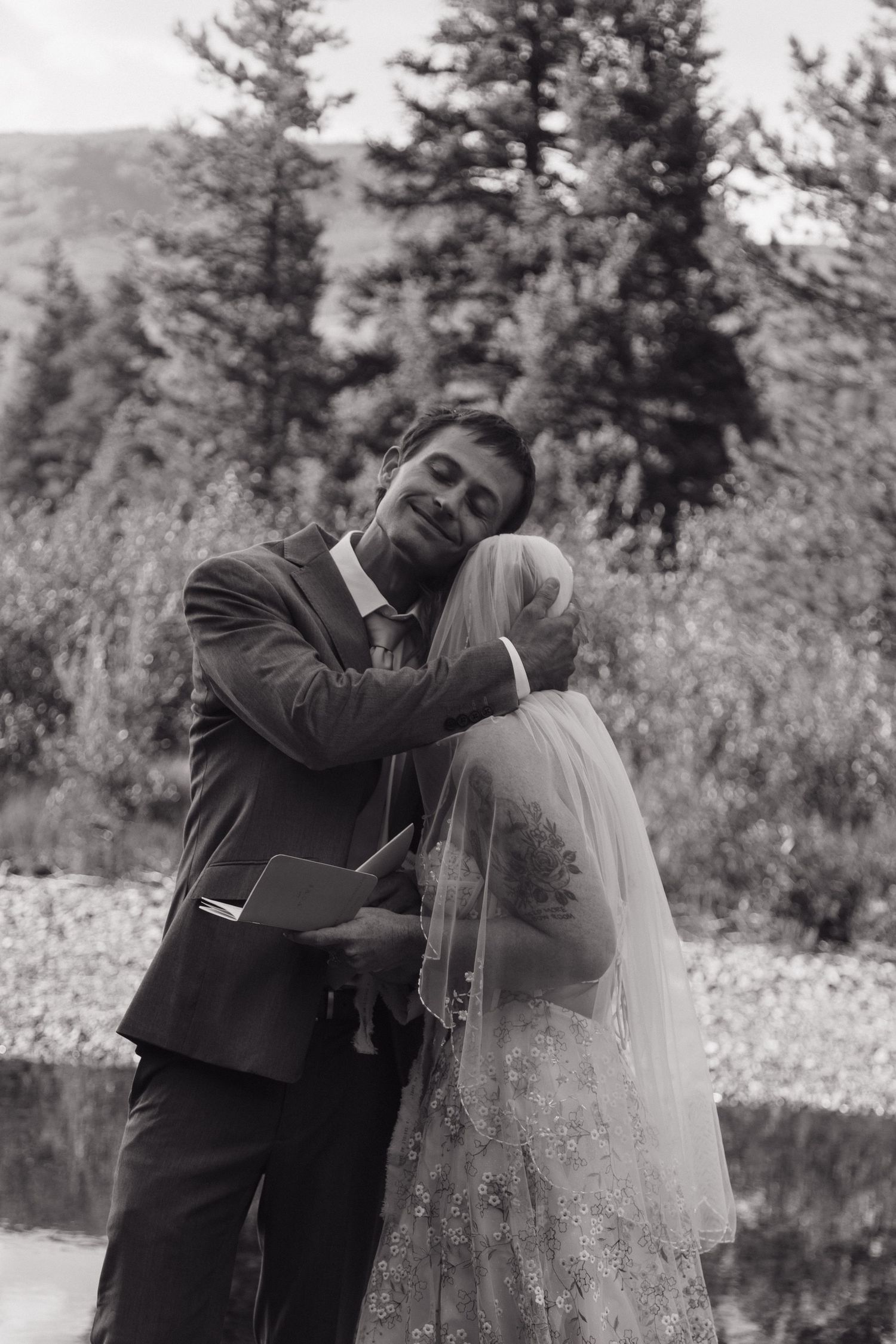 Black and white photo of groom embracing bride in lace veil during outdoor mountain wedding ceremony.