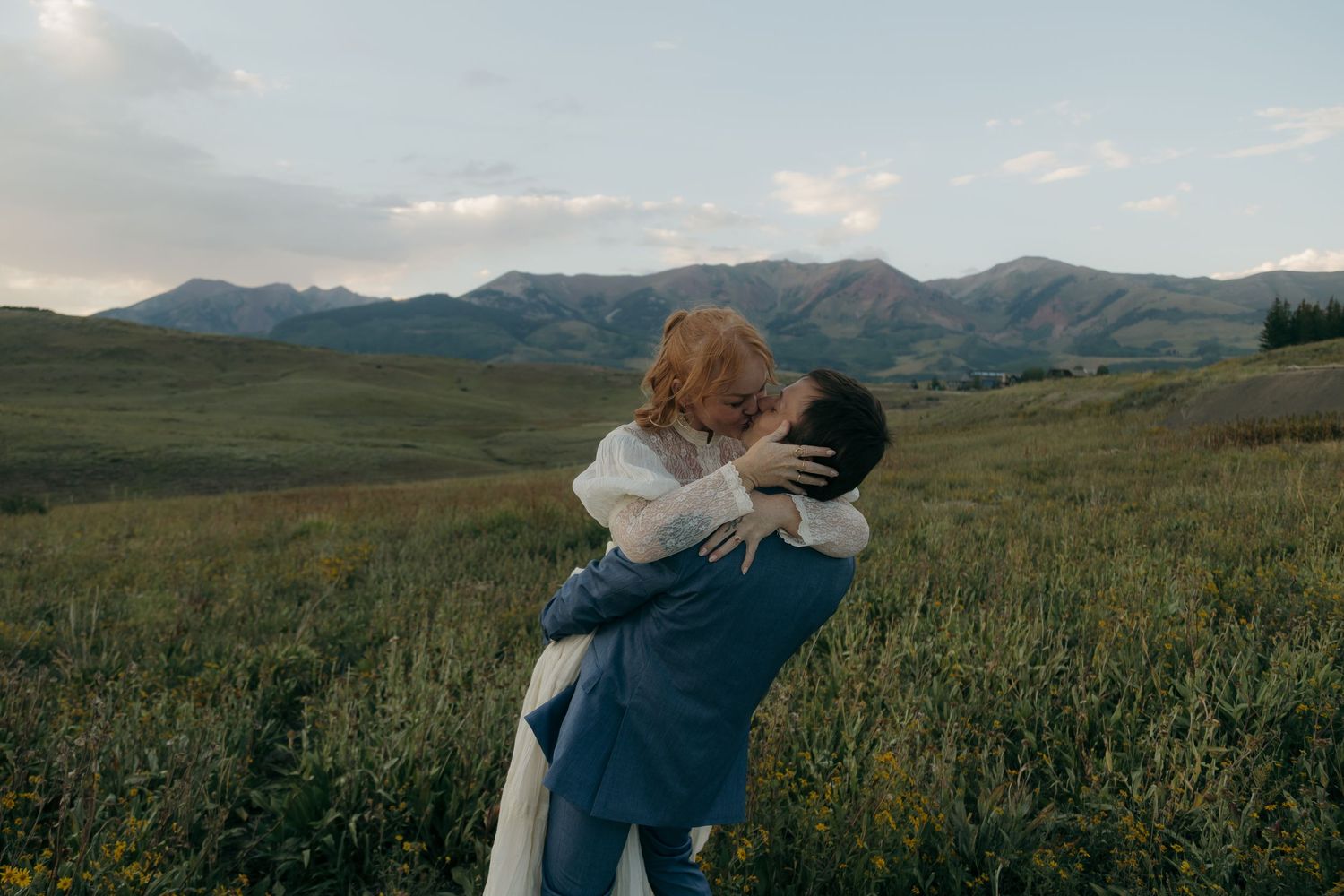 Couple embracing in a lush green meadow with mountain backdrop during golden hour outdoor wedding.