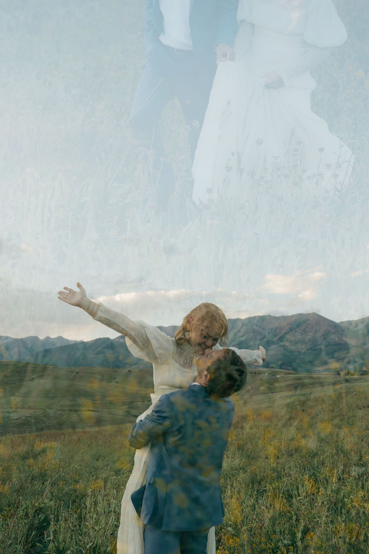 A couple embraces in a green meadow with mountains, woman reaching up, in a dreamy double-exposure wedding photo.