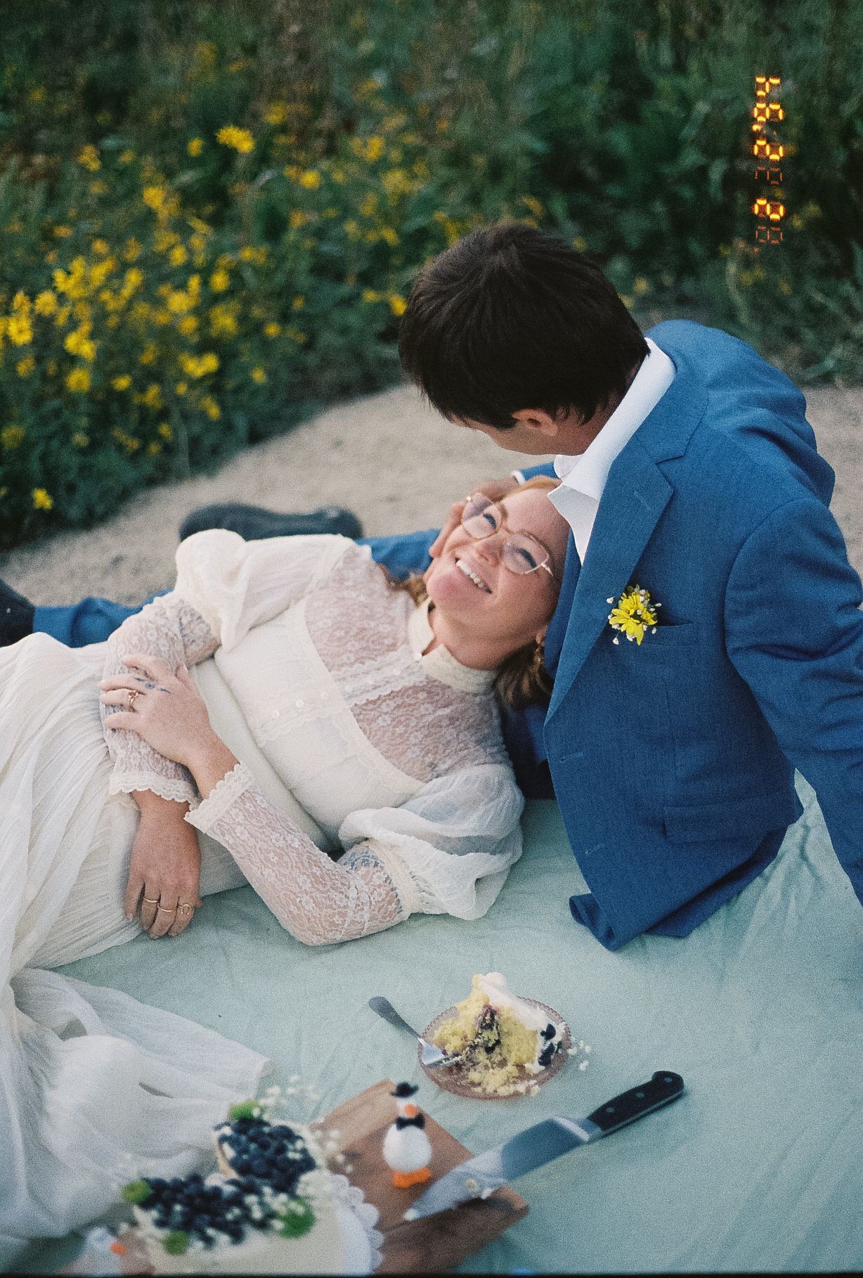 Bride smiling up at groom during romantic outdoor wedding picnic surrounded by yellow wildflowers.