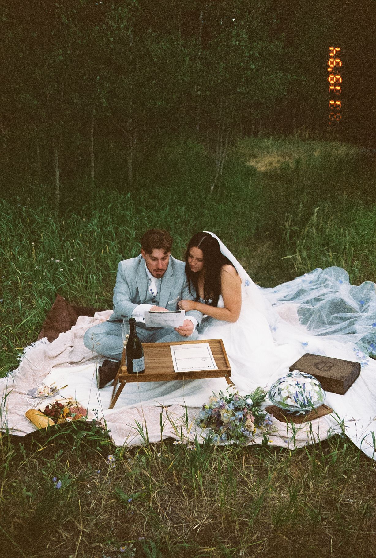 Film photo of bride and groom reading vows together on a white blanket in a grassy field with flowers.
