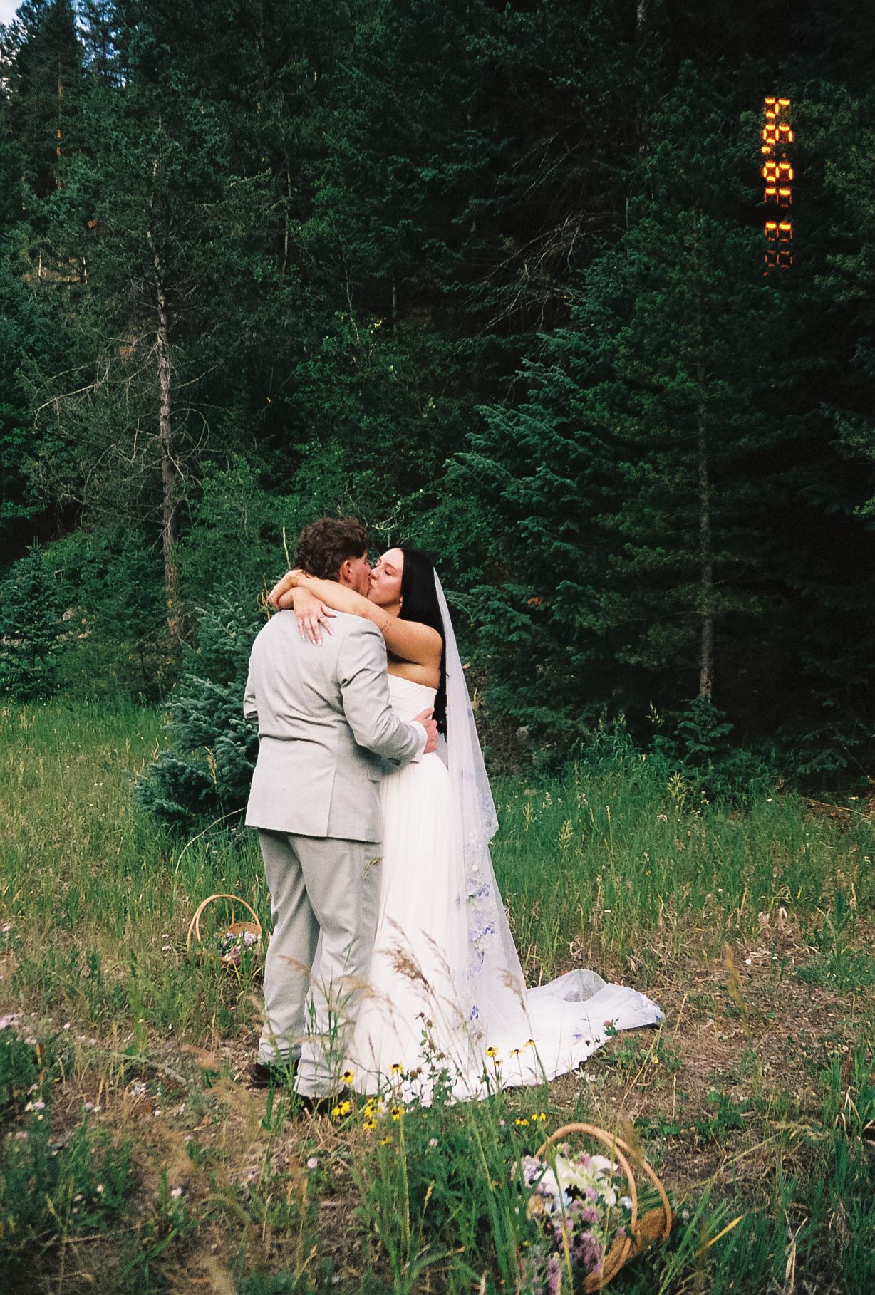 Bride and groom sharing first kiss embrace in wildflower meadow surrounded by evergreen forest trees.