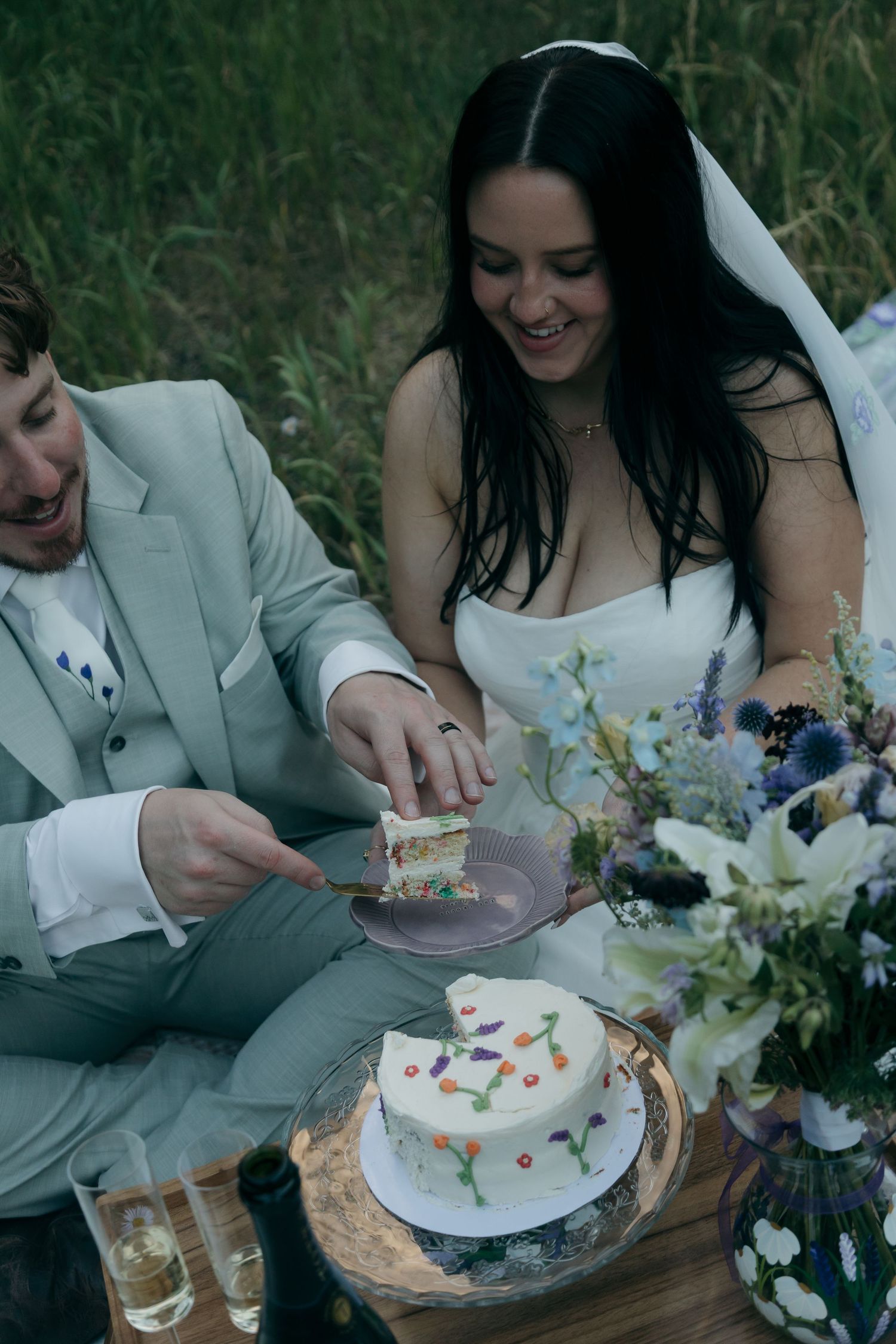 Bride and groom cutting their white wedding cake with colorful decorations during an outdoor picnic celebration.