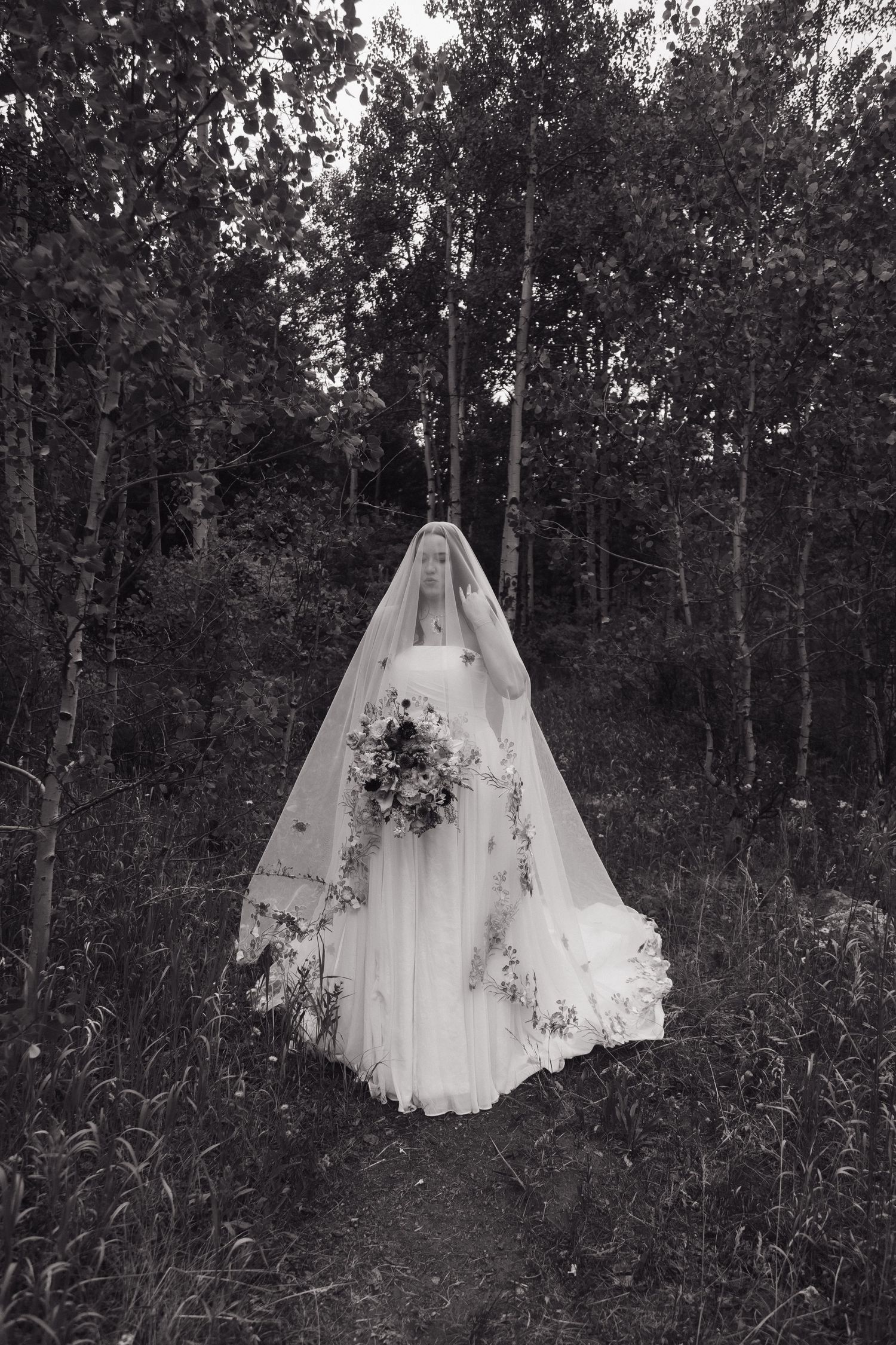 Black and white photo of a bride in a long veil and gown holding a bouquet, standing on a forest path.