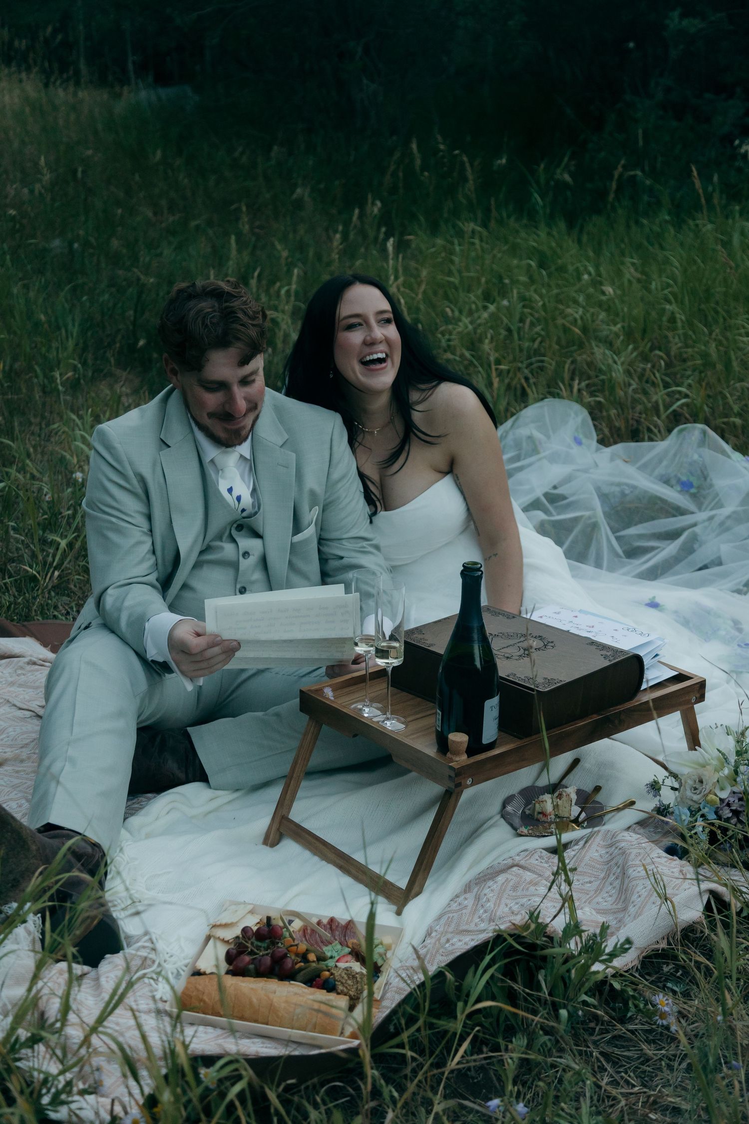 Joyful bride and groom reading together during an elegant outdoor picnic with wine and charcuterie in nature.