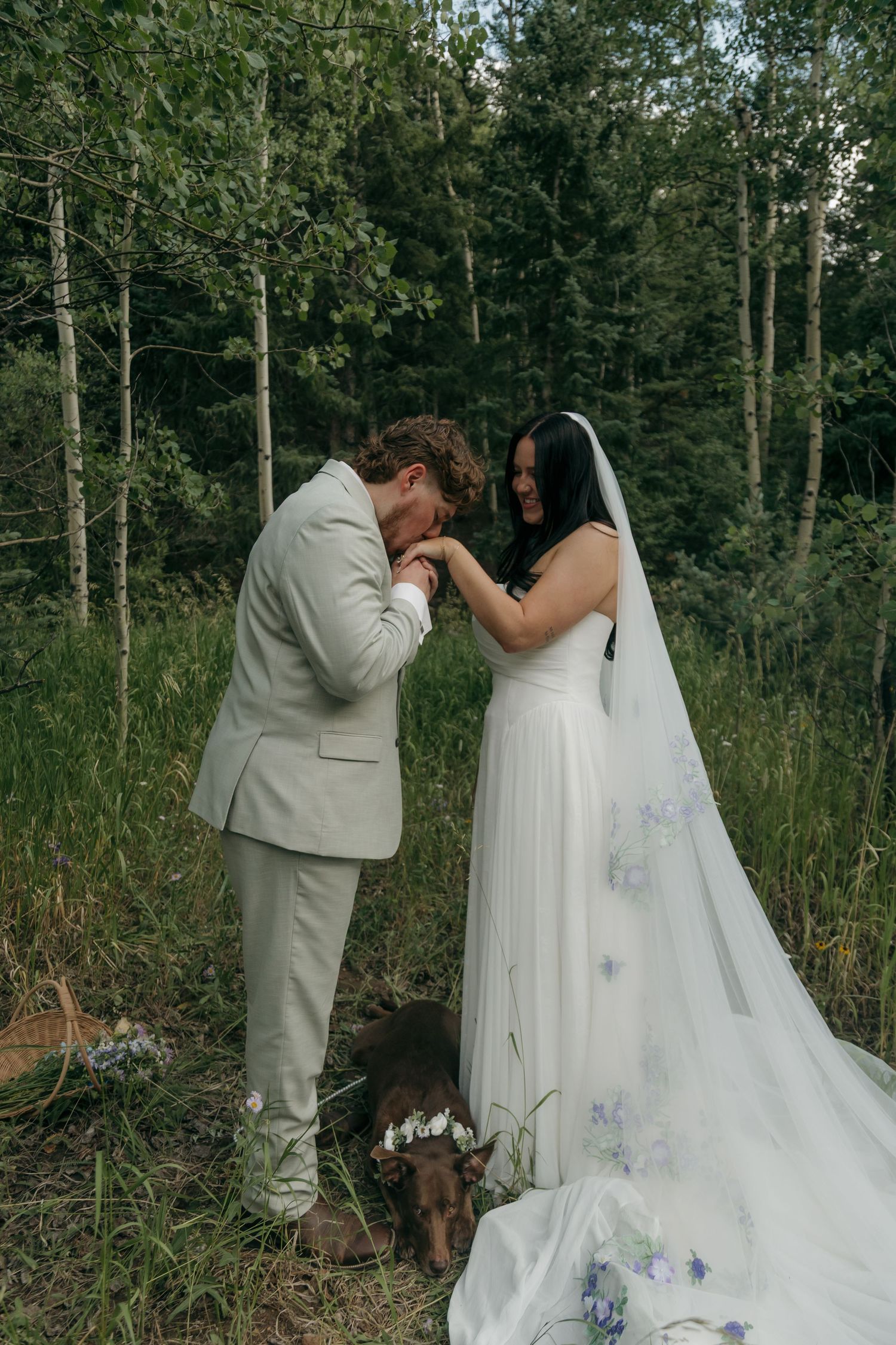 Groom gently kisses the hand of his smiling bride as their dog wears a floral crown at their feet.