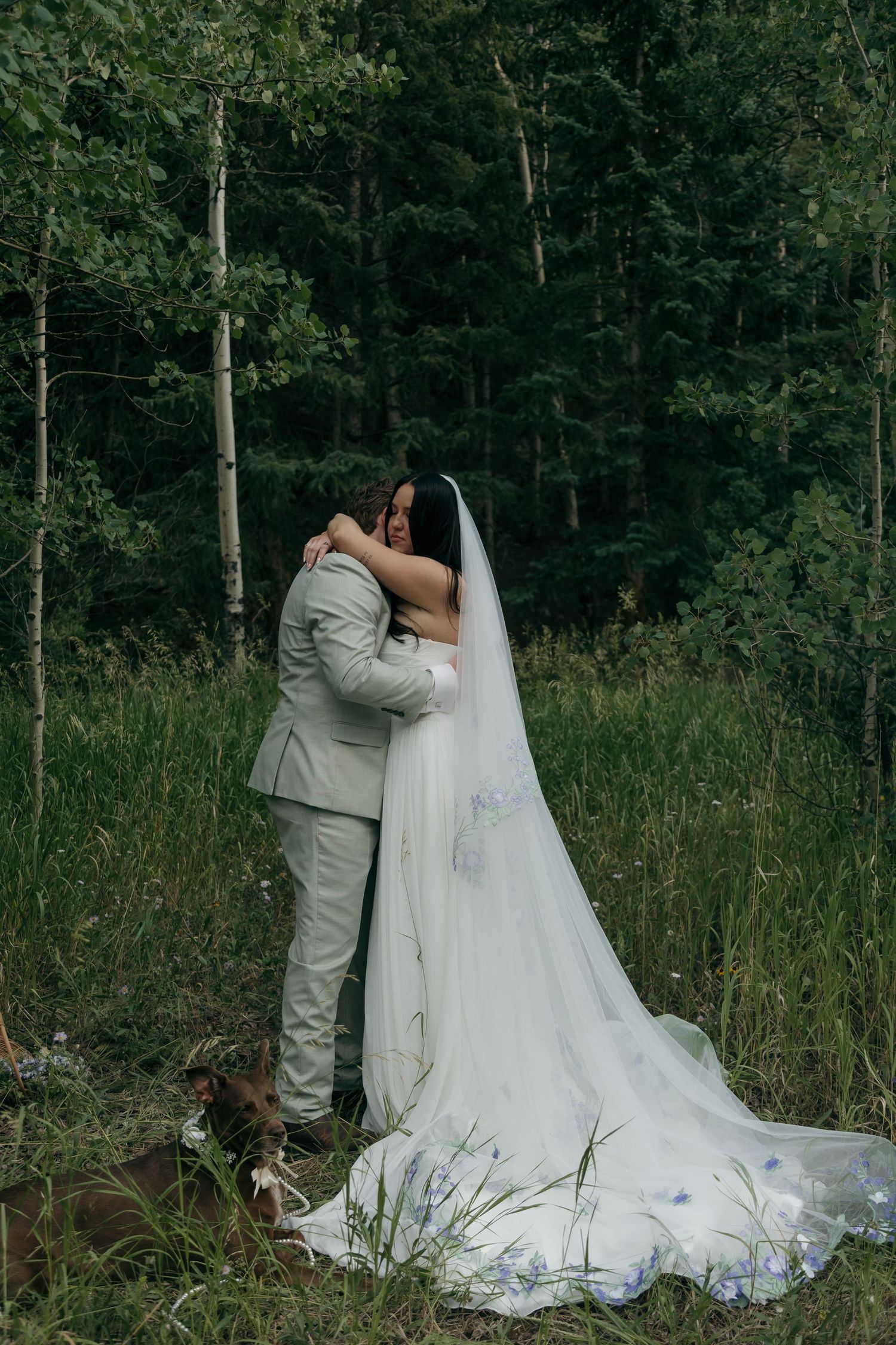 Newlyweds share first embrace after forest wedding ceremony, bride in floral embroidered gown and veil.