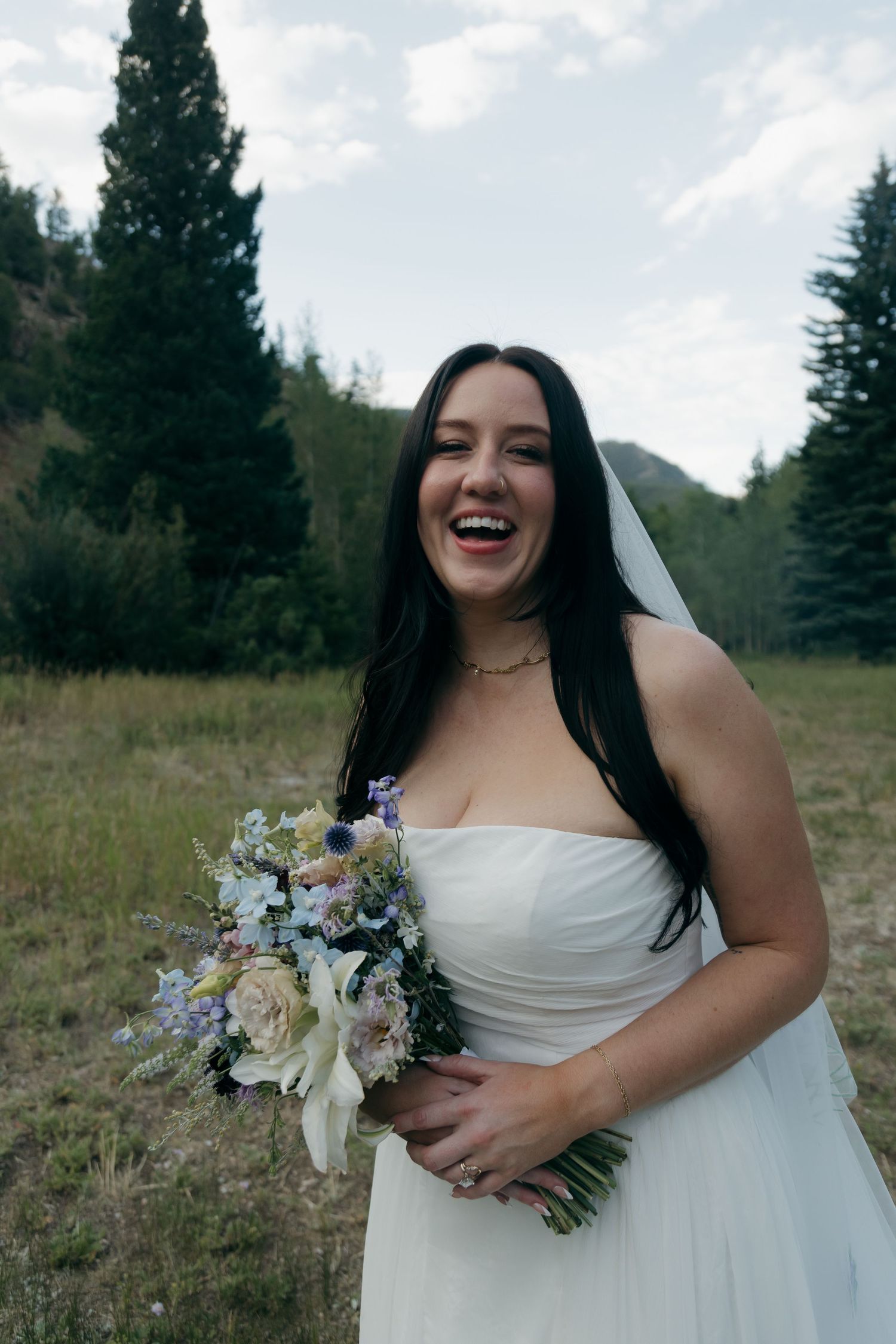Laughing bride with long dark hair holds a pastel wildflower bouquet in a strapless white gown against a mountain backdrop.