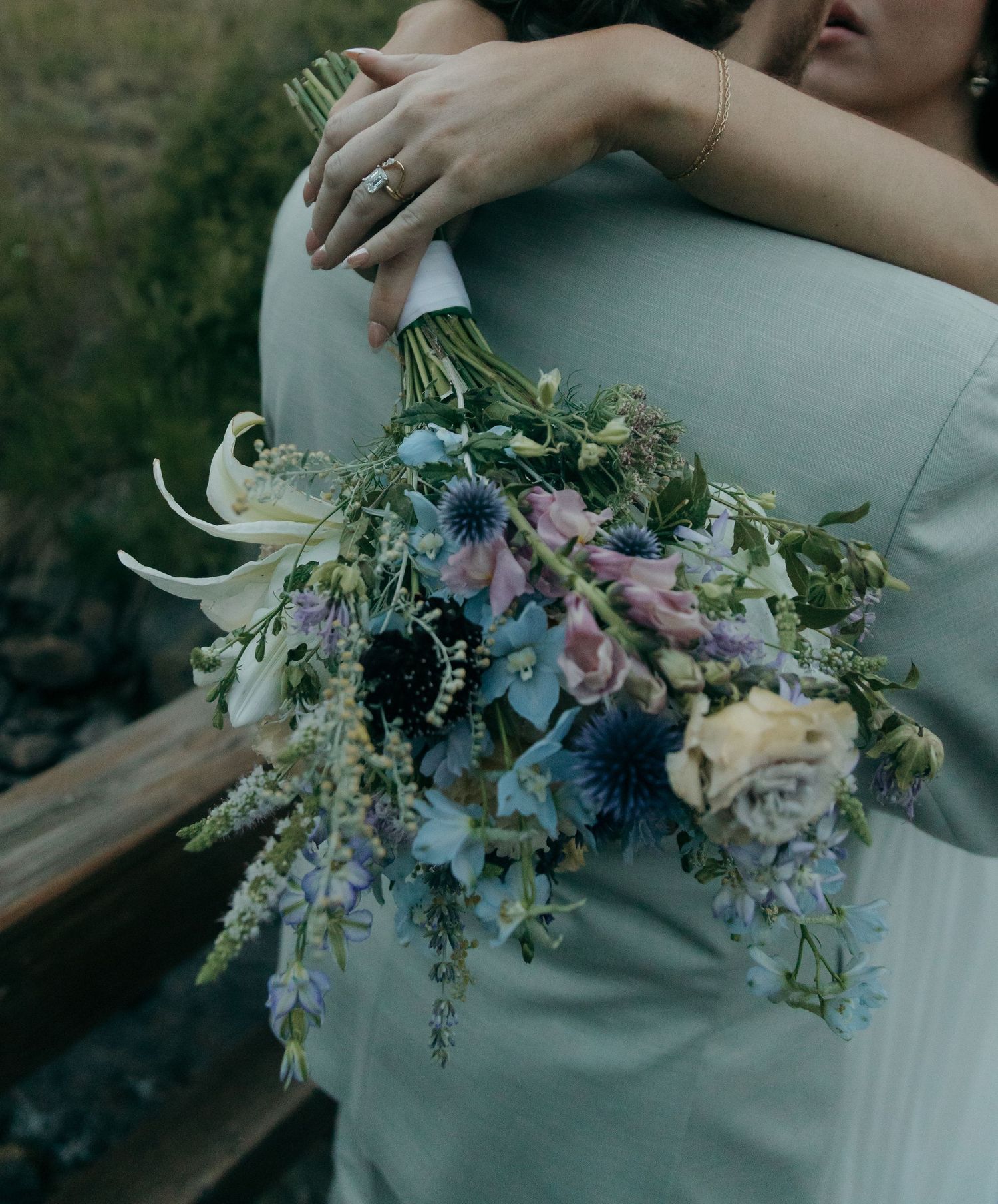 Bride embracing groom from behind while holding cascading blue, purple, and white wildflower wedding bouquet outdoors.
