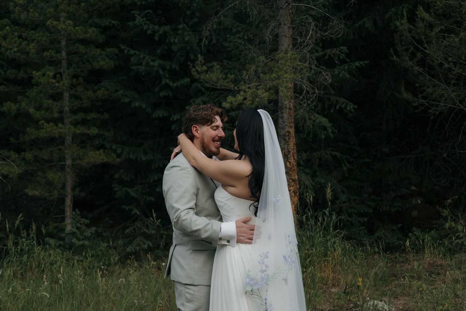 Laughing bride and groom share a joyful embrace in a green forest setting.