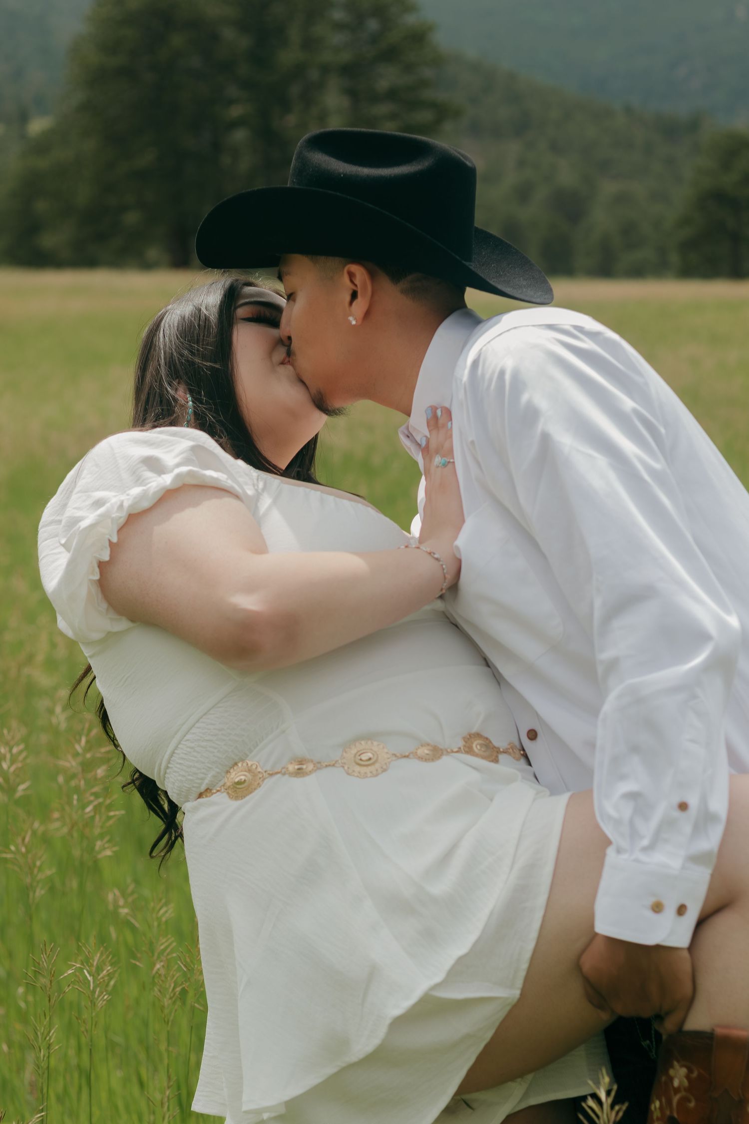 Intimate moment between couple in cowboy attire amidst tall grass with mountains in background.
