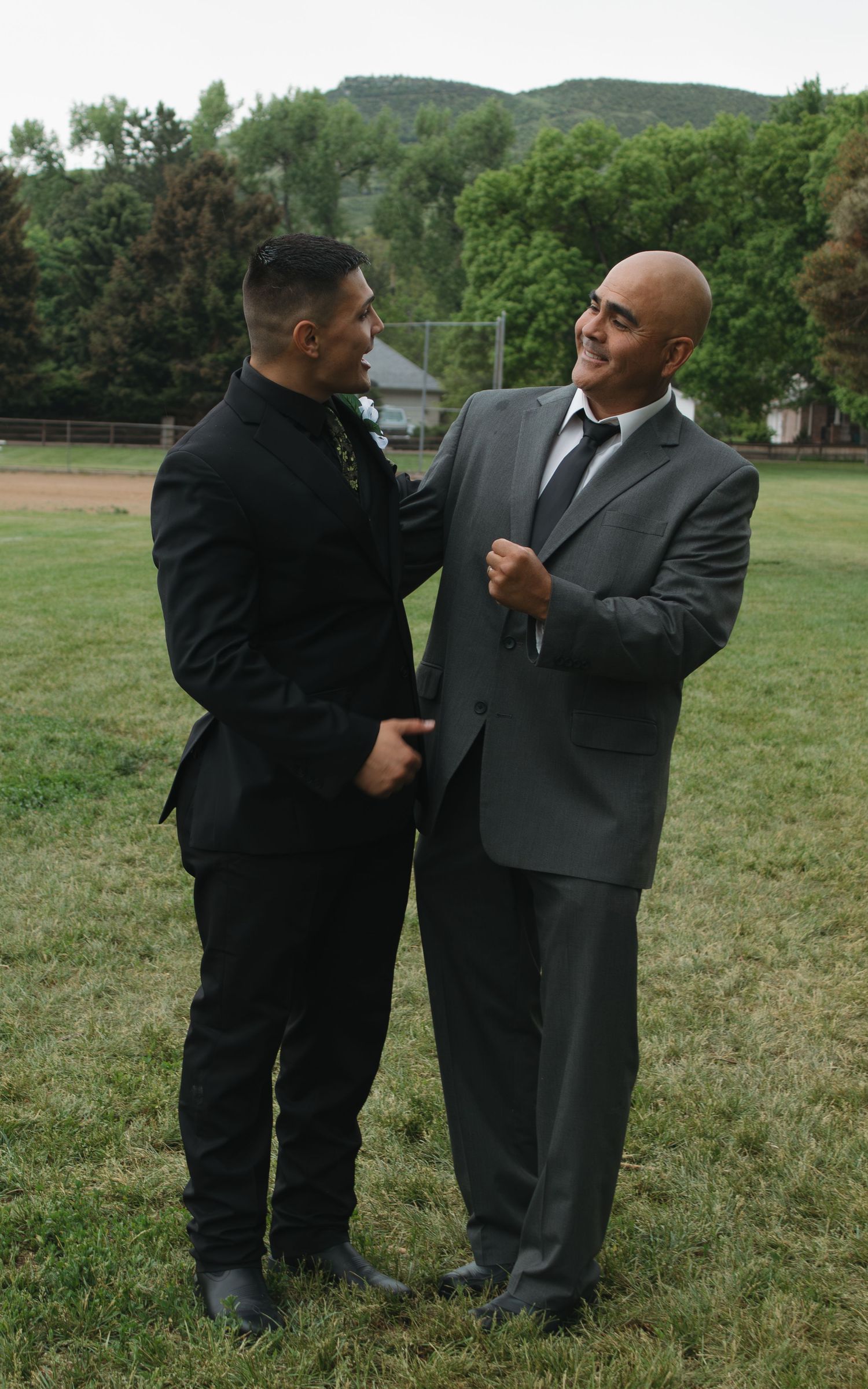 Two men in formal suits share a moment on a grassy lawn with mountains in the background.