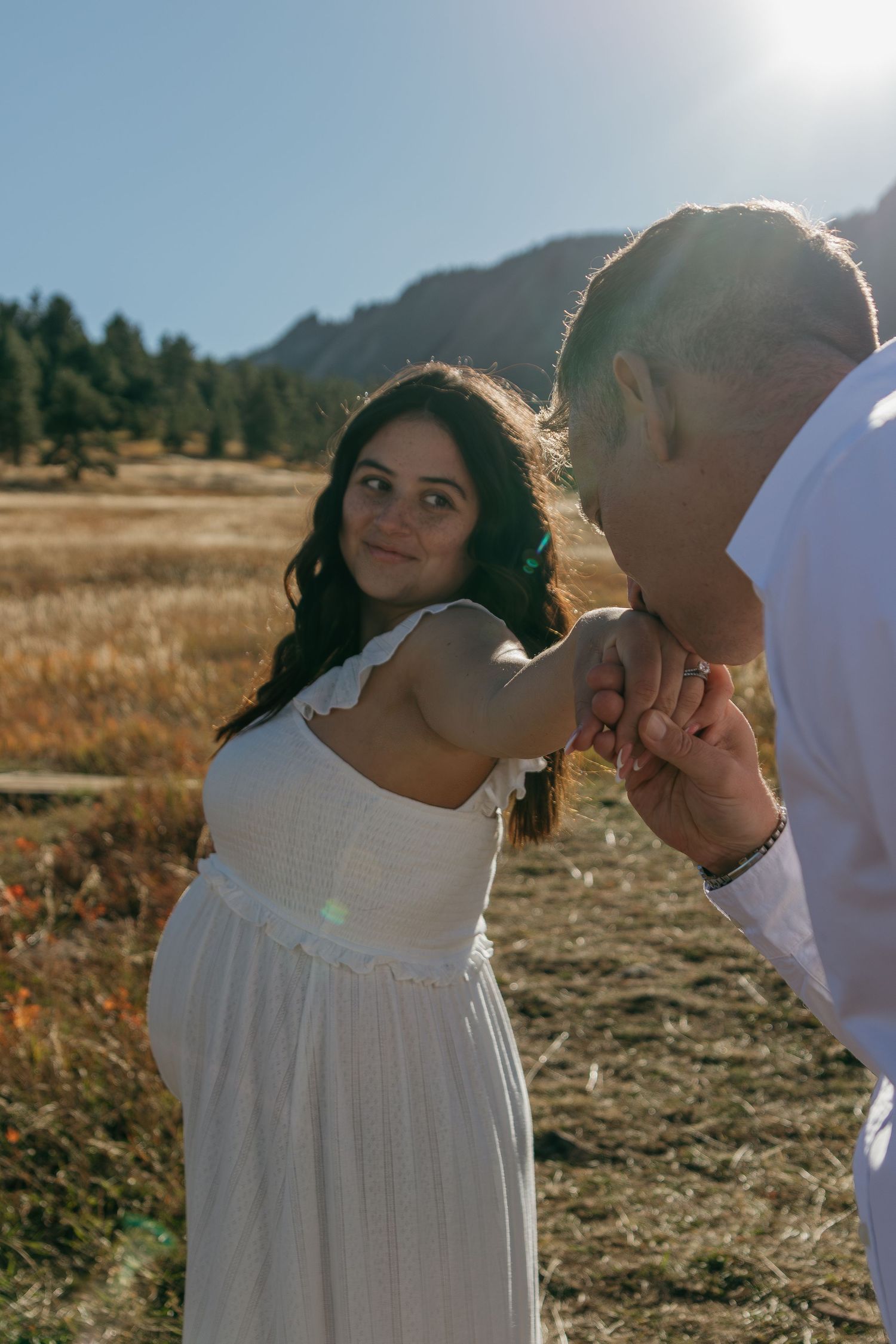 A couple shares an intimate moment in a golden field during sunset, with mountains visible in the background.