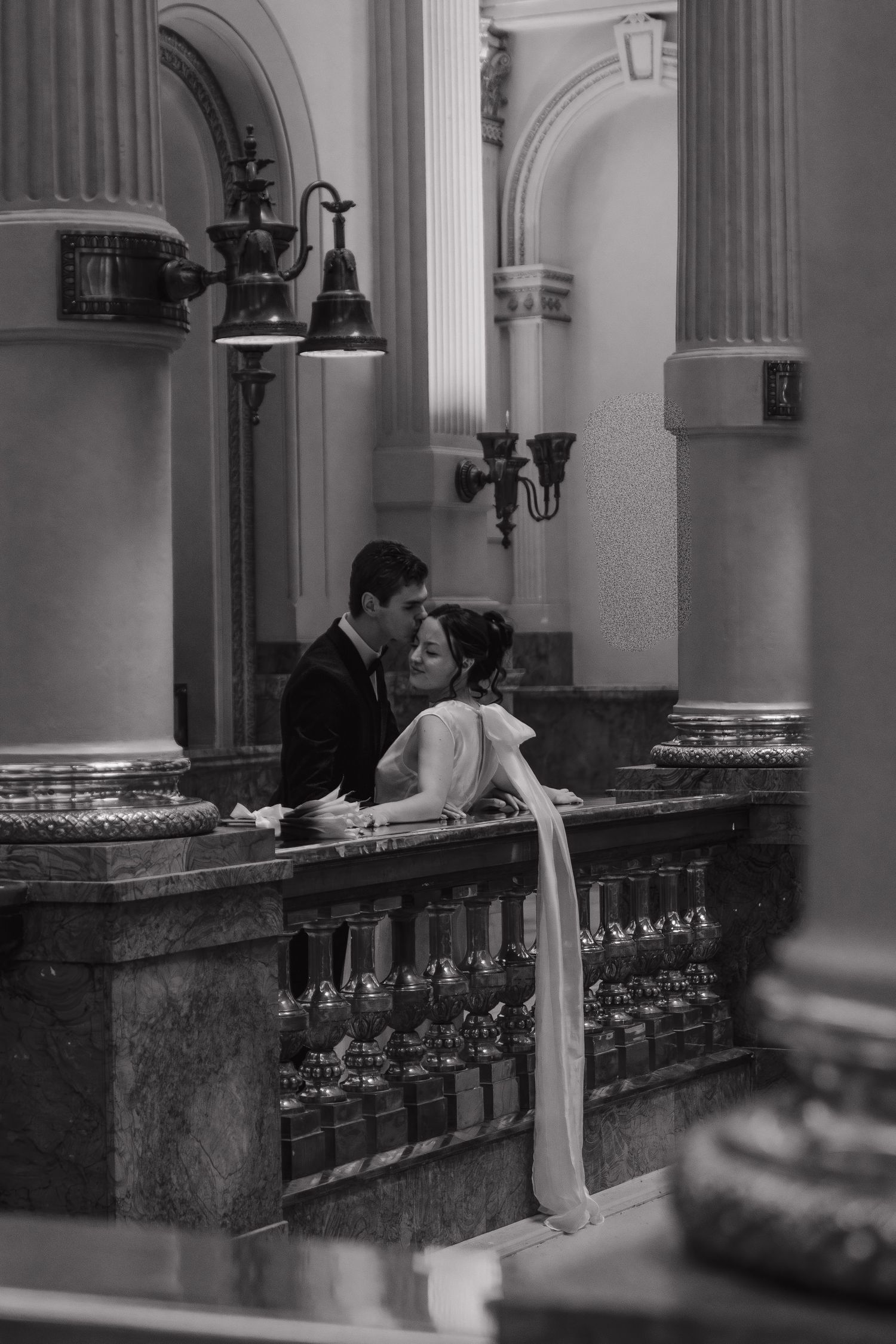 Black and white photo of couple by ornate marble railing in classical interior.