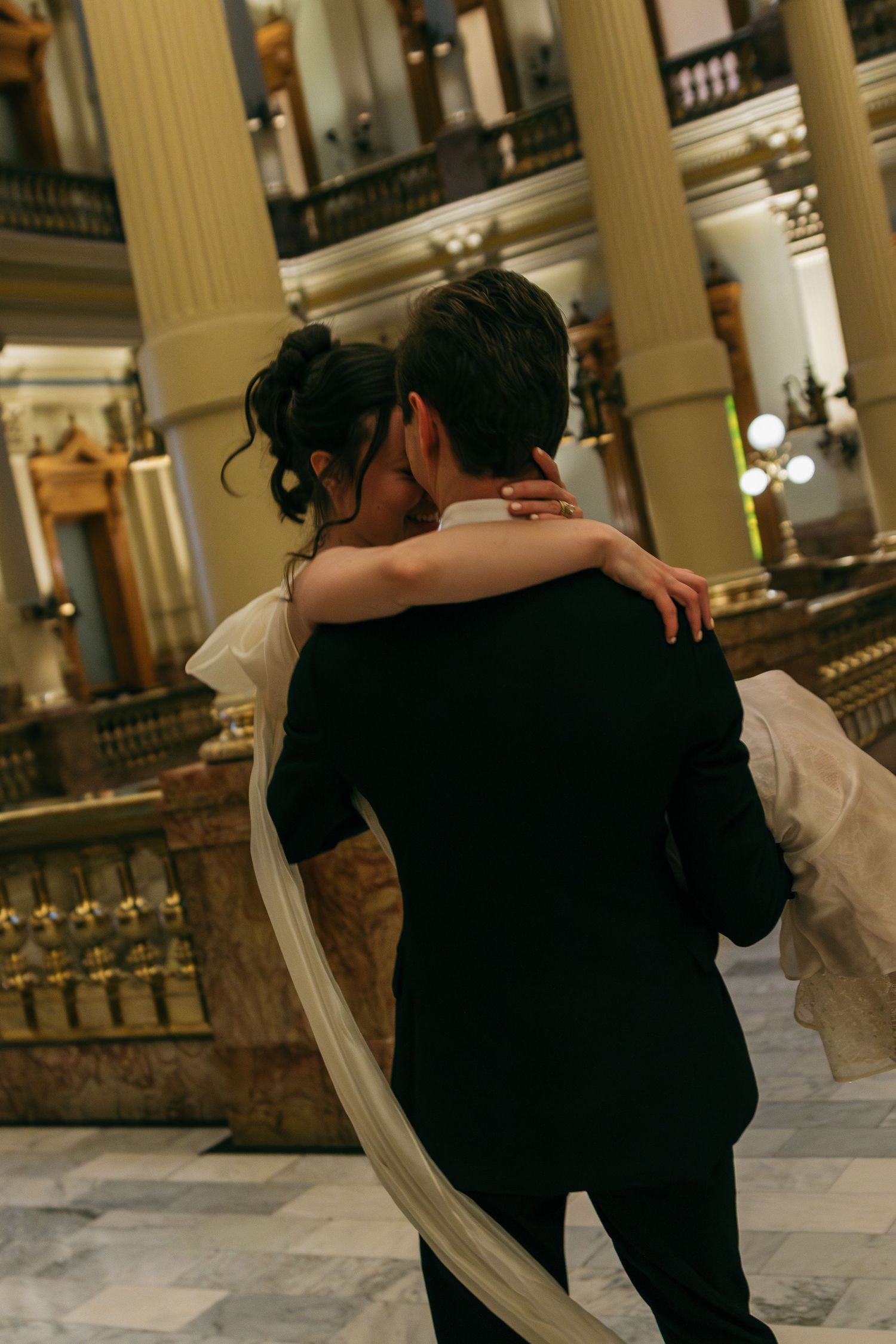 A couple embraces in an elegant venue with classical columns and marble floors.