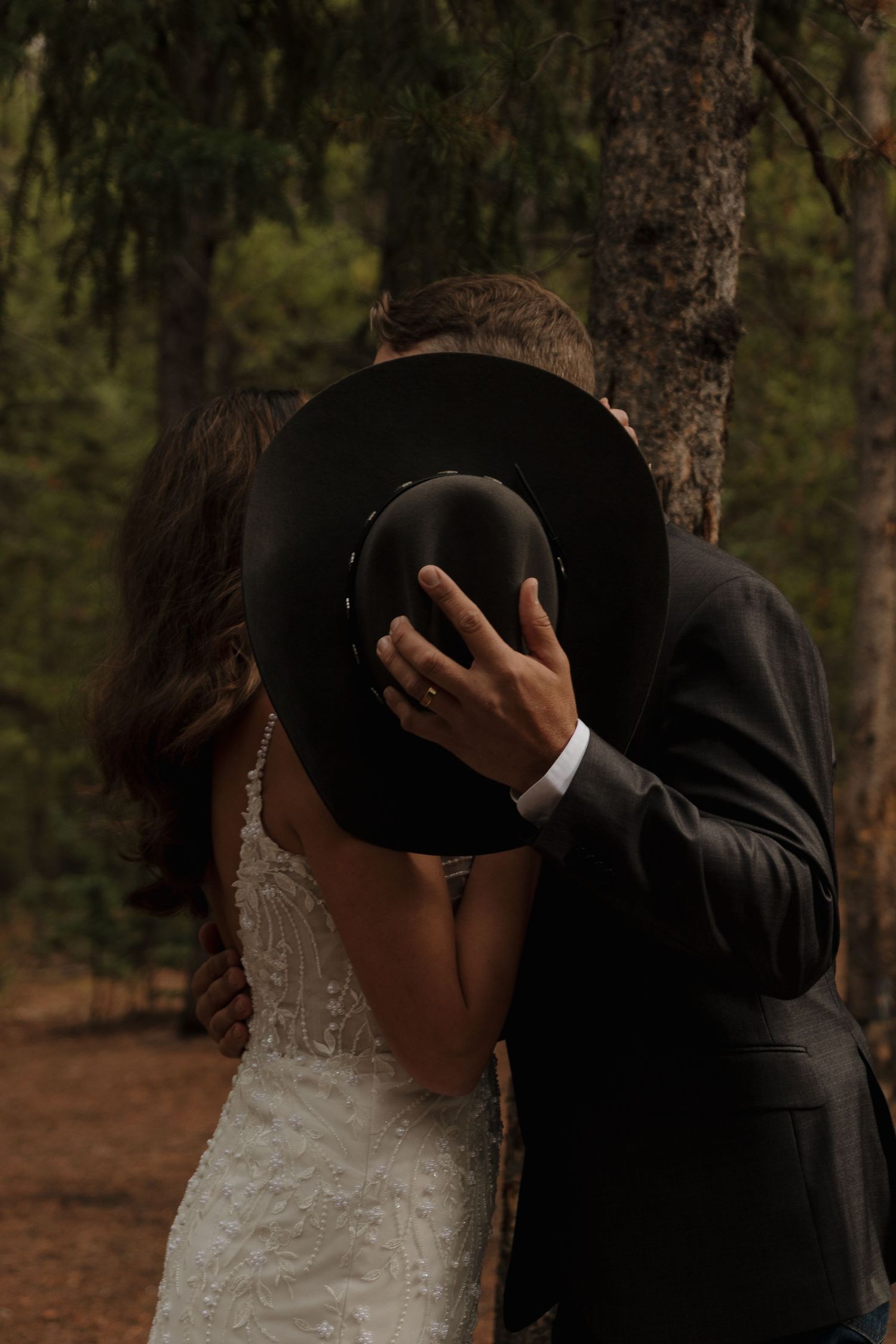 A romantic moment shared behind a black cowboy hat in a forest setting.