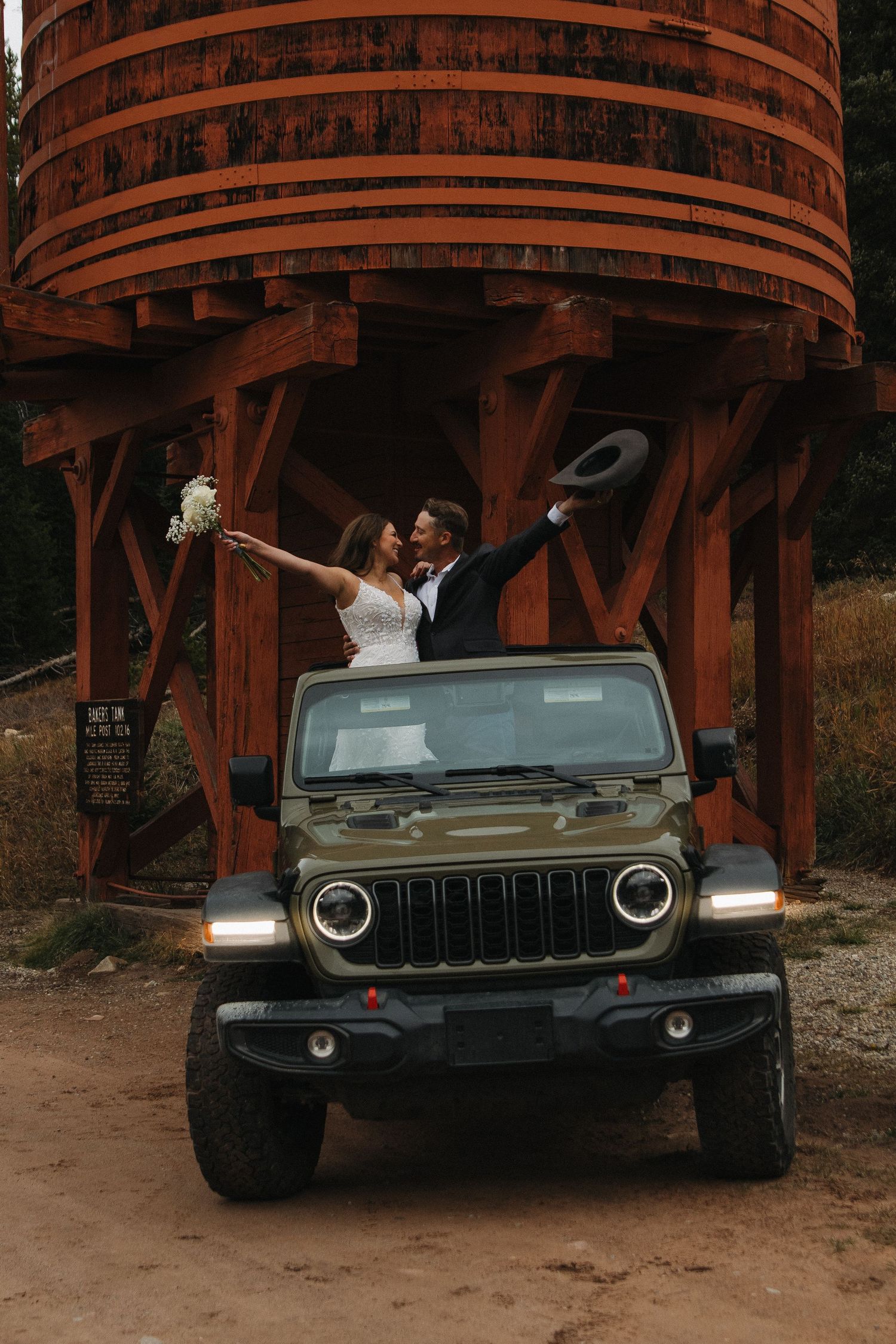 Newlyweds celebrate standing through a Jeep sunroof under a rustic wooden structure.