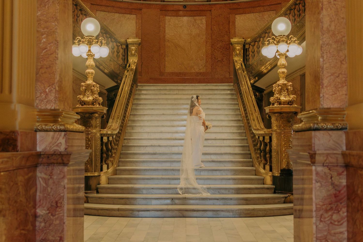 A bride in a white wedding gown stands on an elegant marble staircase in a luxurious palace setting.