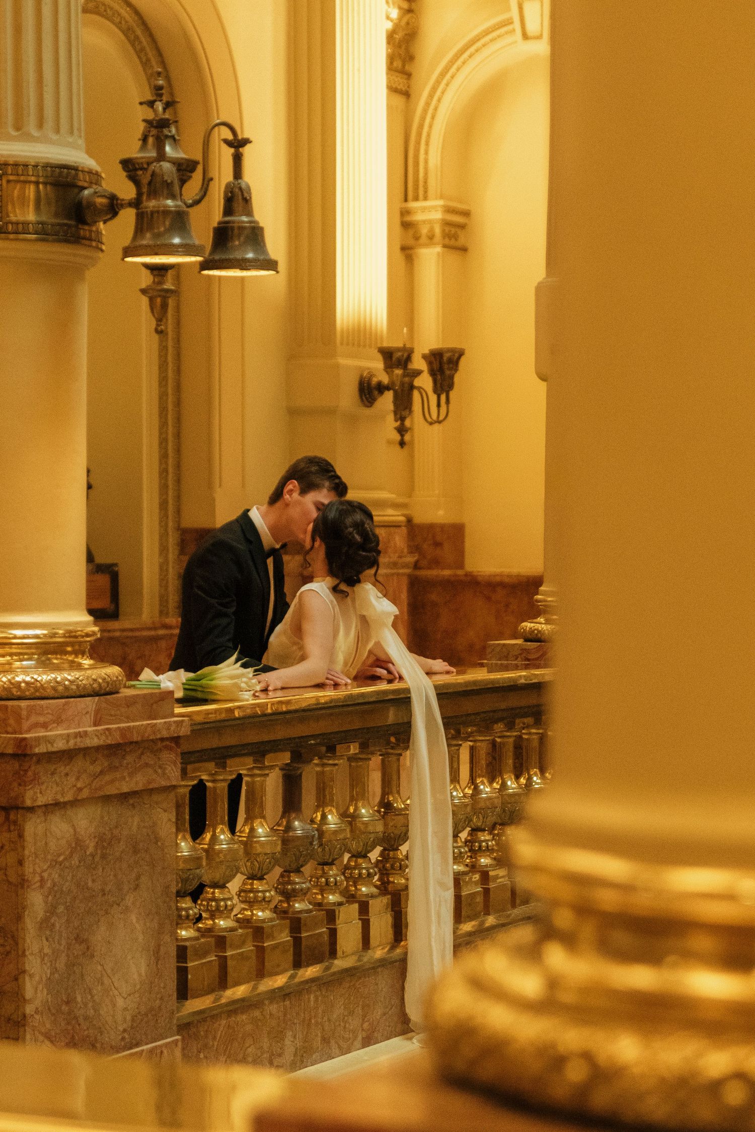 A romantic couple leans over an ornate golden balustrade in a grand historic building with warm lighting.