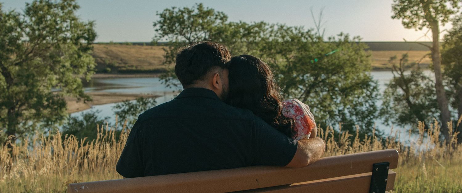 A couple sits together on a bench overlooking a serene lake at sunset.