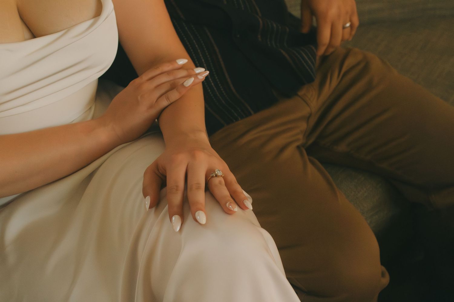 Close-up of hands with white nail polish resting on white and brown fabric with warm lighting.