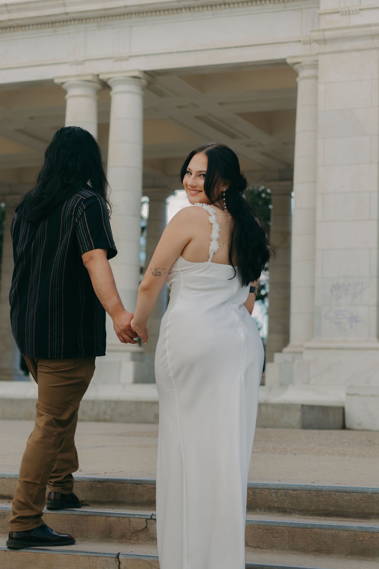 A couple in formal attire stands on marble steps near classical columns of a grand building.