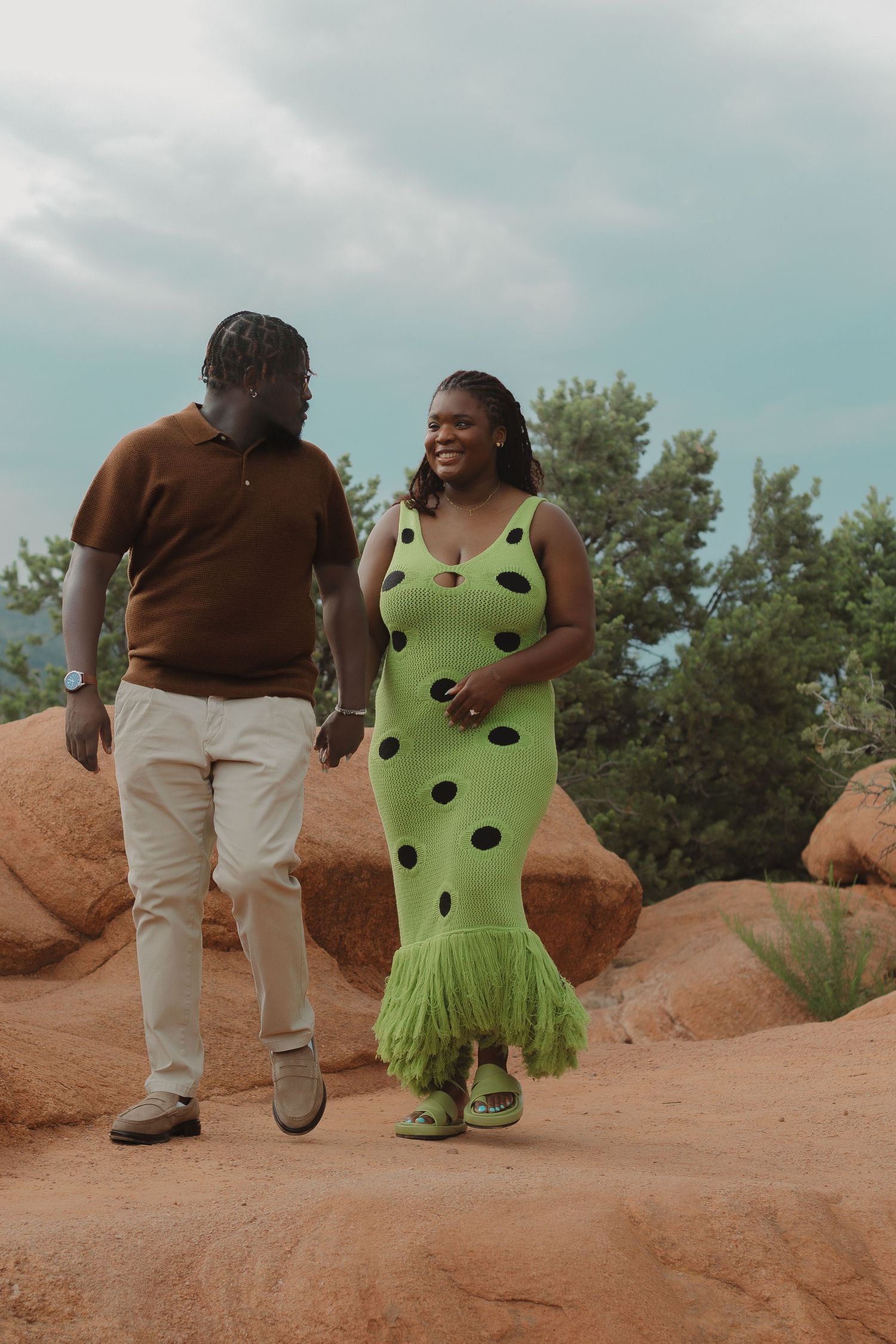 A couple walks together on red rocks wearing a brown sweater and lime green polka dot dress with fringe detail.