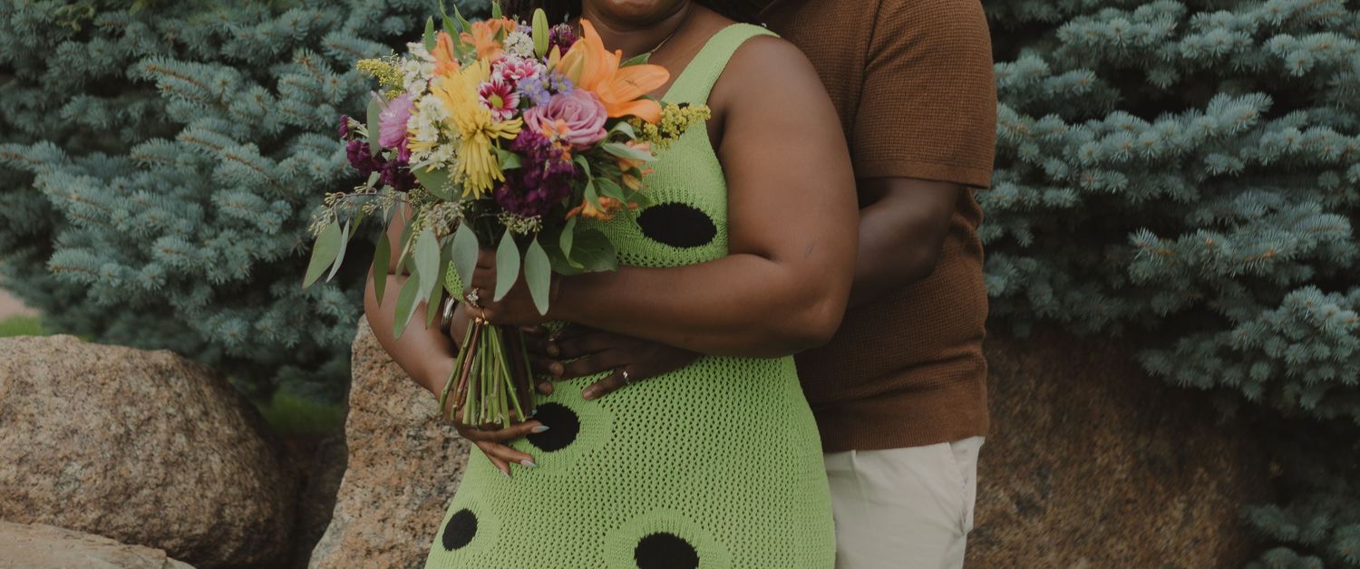 A person in a green polka dot dress holding a colorful bouquet of flowers stands against a backdrop of blue spruce trees.