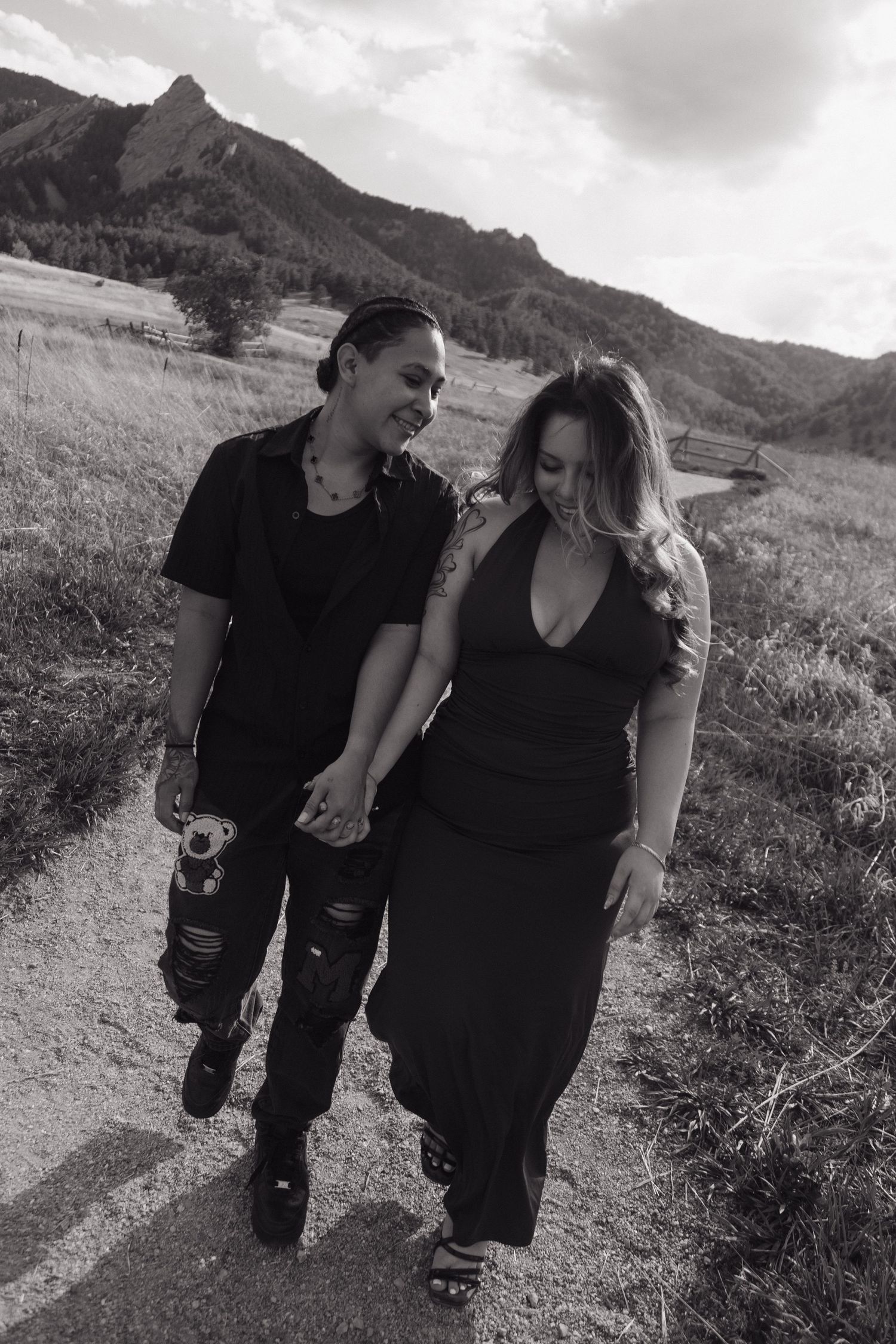 Couple in black outfits walk hand-in-hand on a mountain path with rocky flatirons in the background.