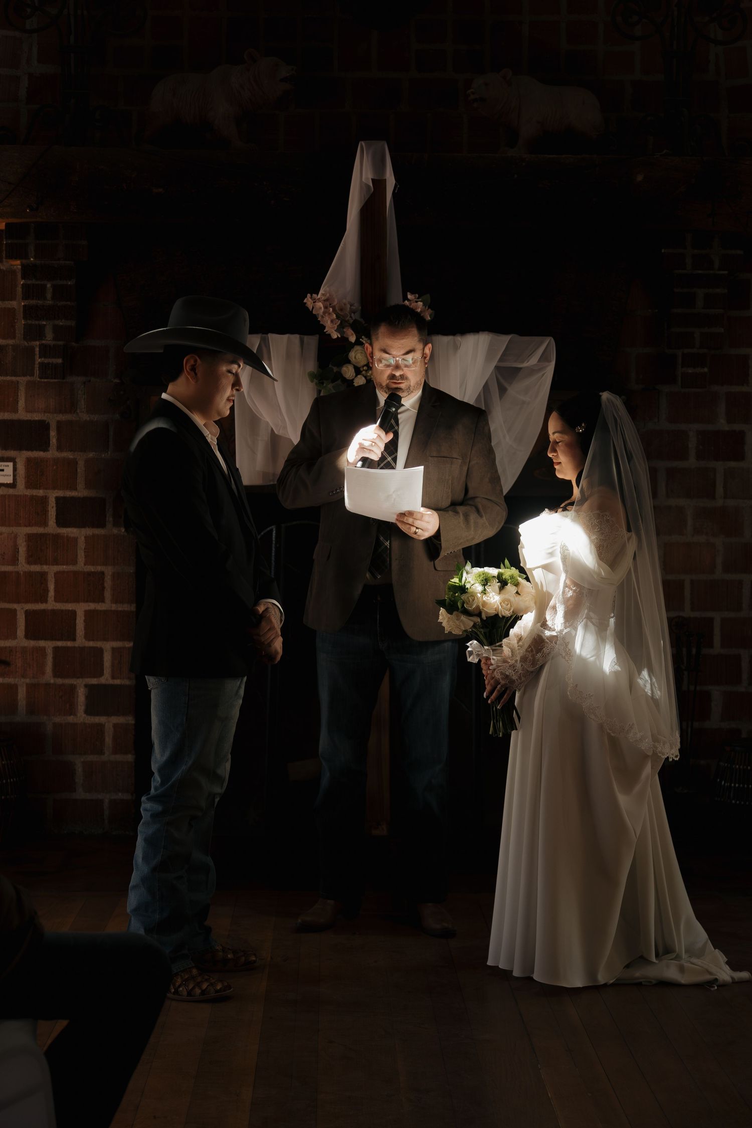 Bride and groom exchange vows in dimly lit brick venue with officiant holding illuminated paper.