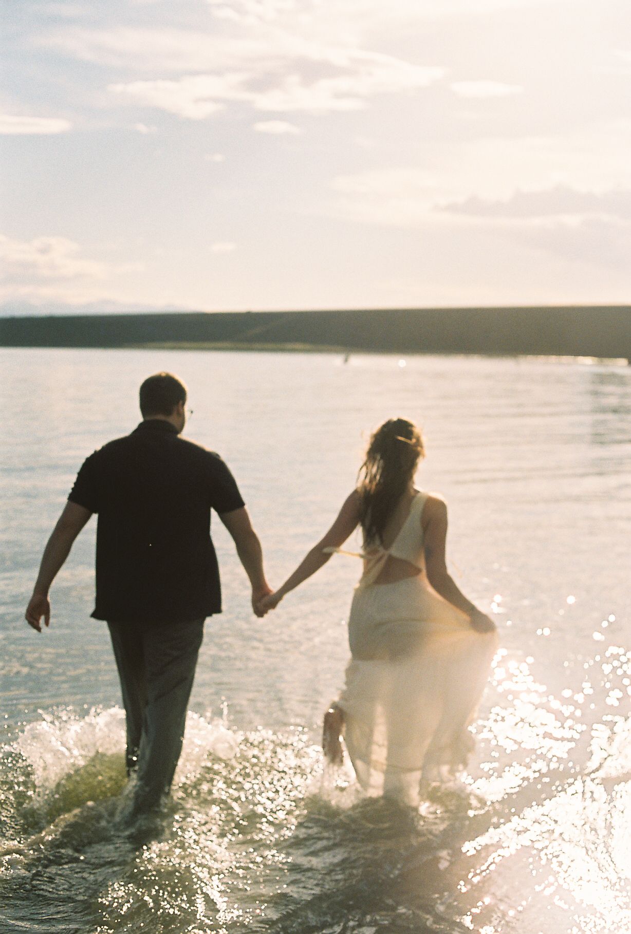 A couple holds hands while wading through sparkling lake water at sunset, walking away from the camera together.