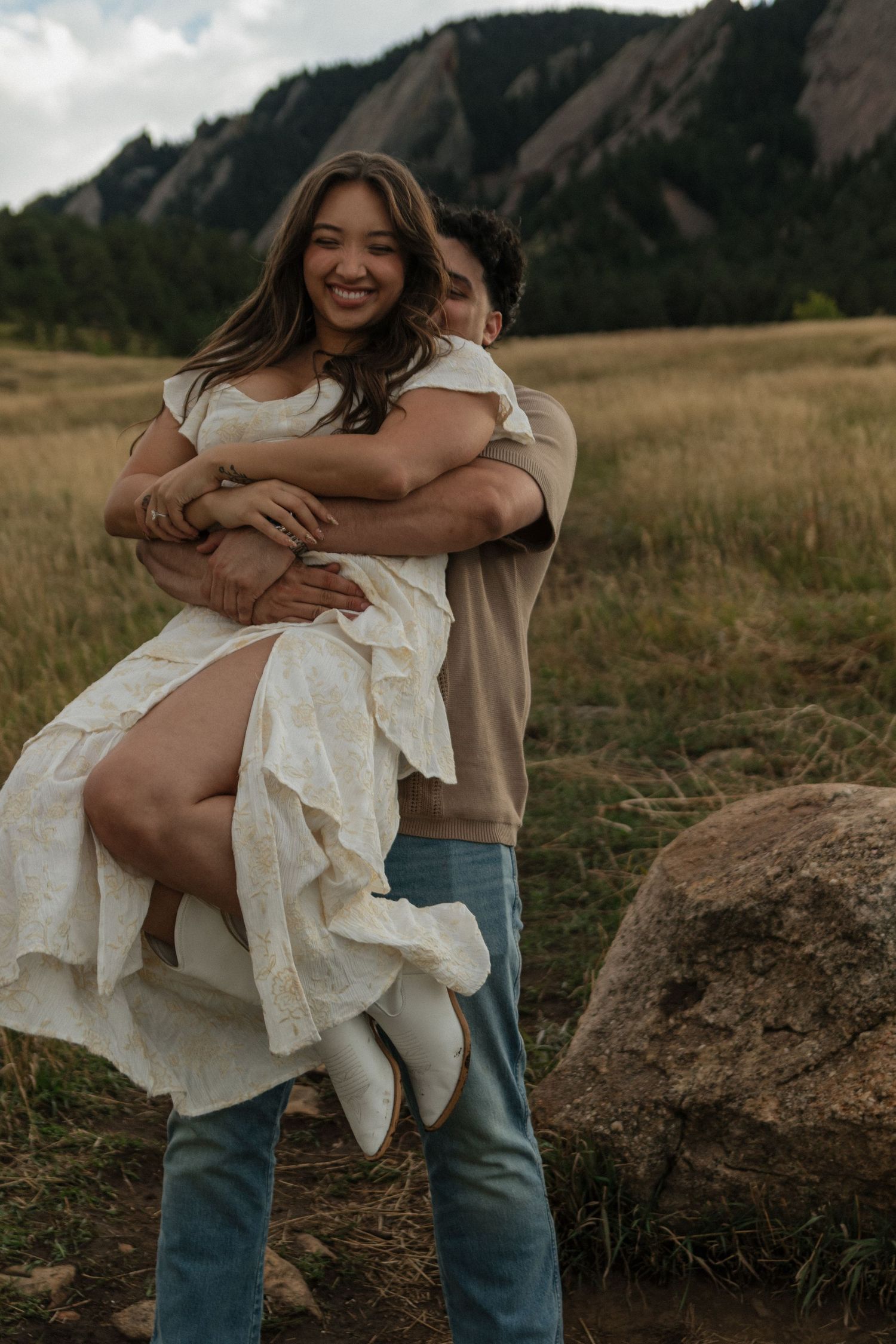Man lifting smiling woman in white dress in golden meadow with Flatirons backdrop.