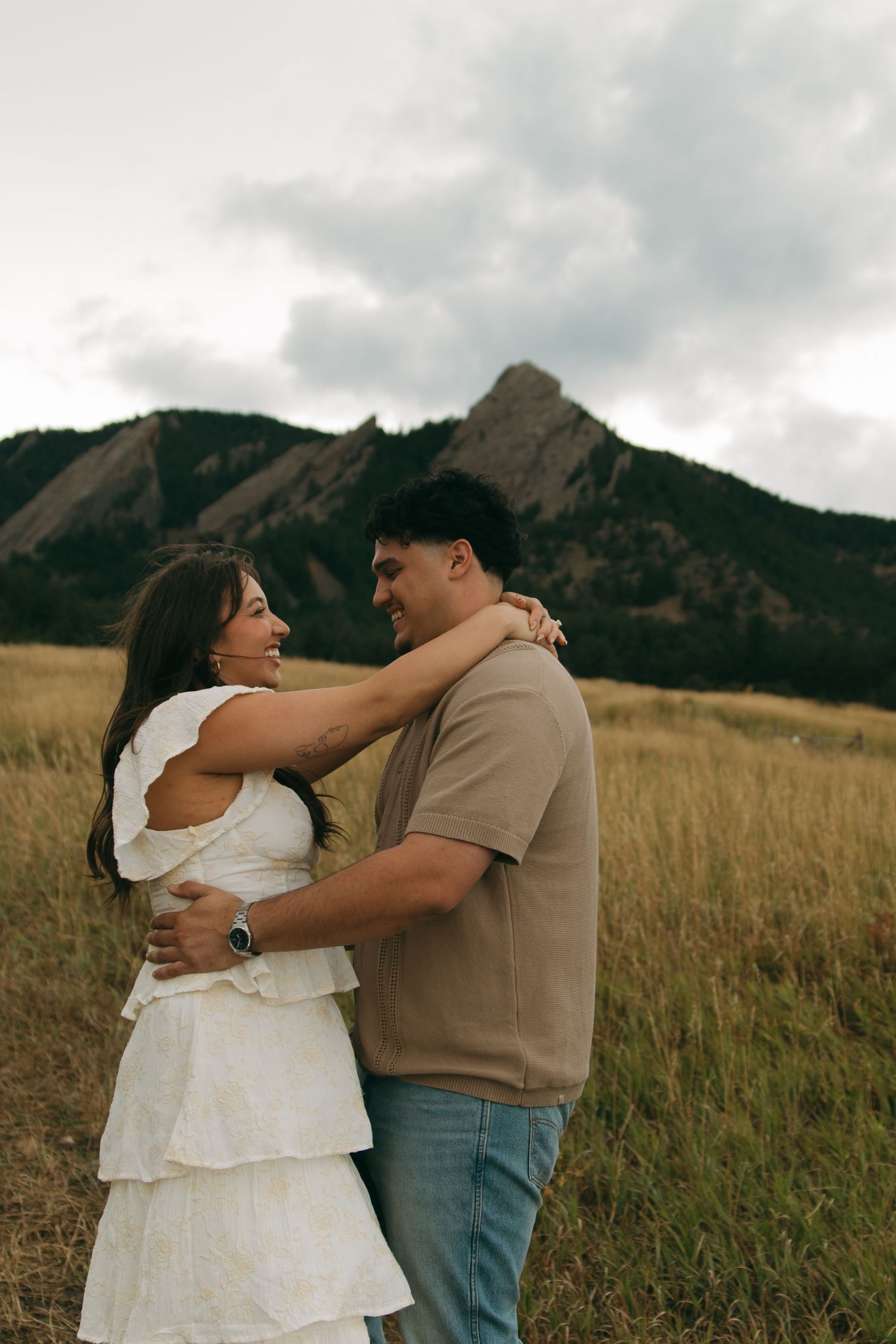 Smiling couple embracing in golden grass field with rocky Flatiron mountains behind them.