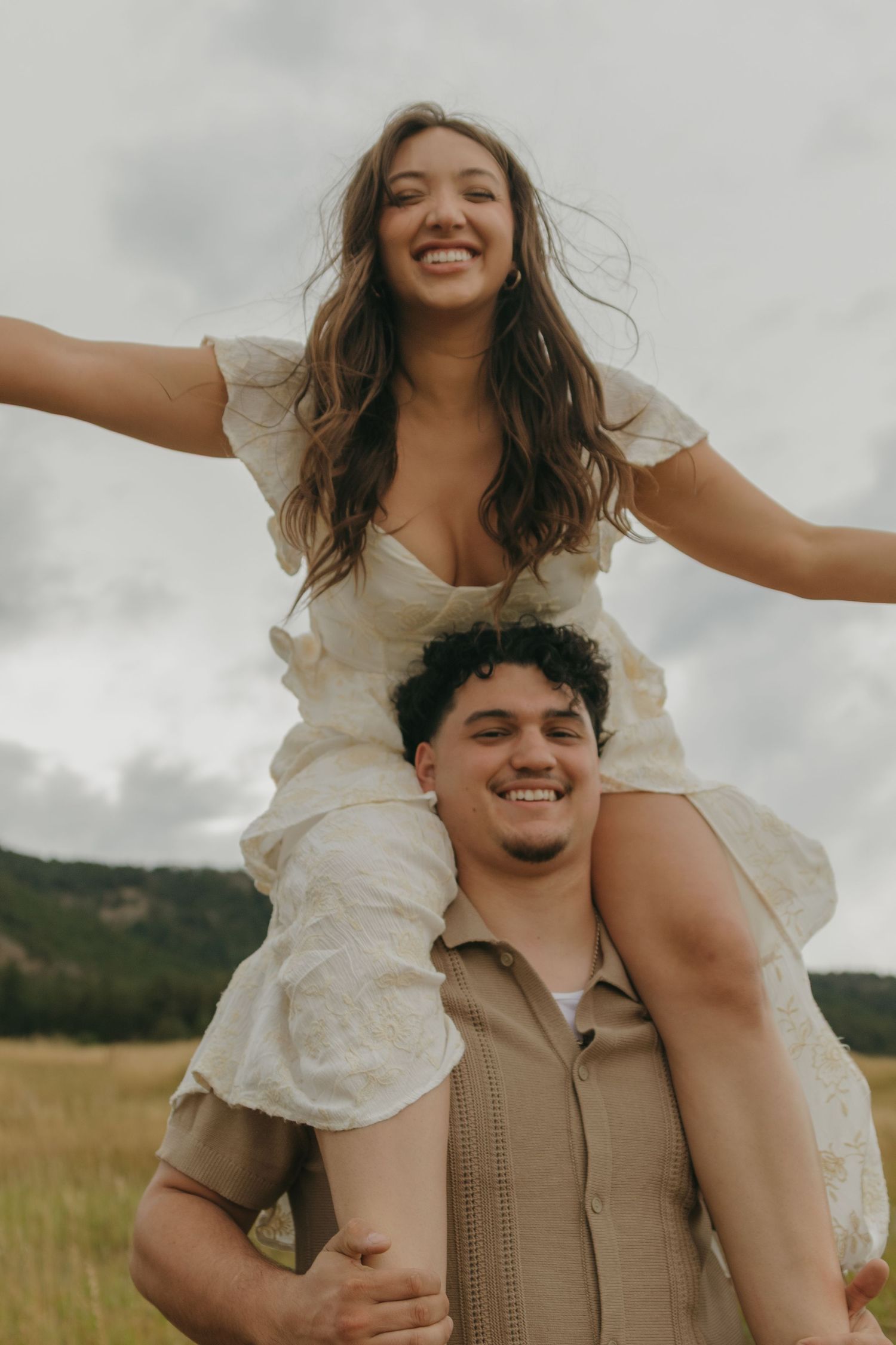 Joyful woman in white dress sits on man's shoulders with arms outstretched in a golden field.