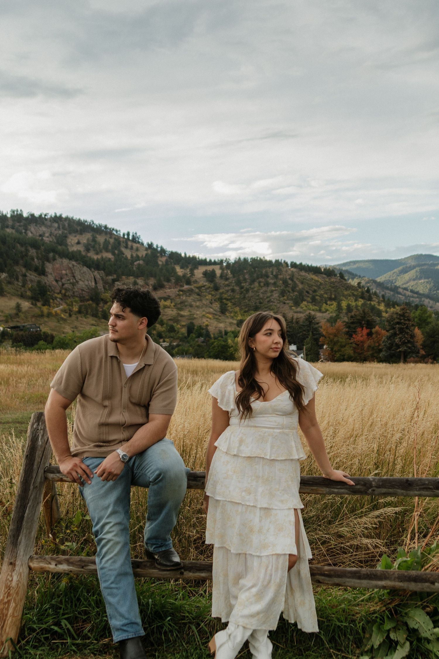 Couple poses by rustic fence in golden field with mountain backdrop on cloudy day.