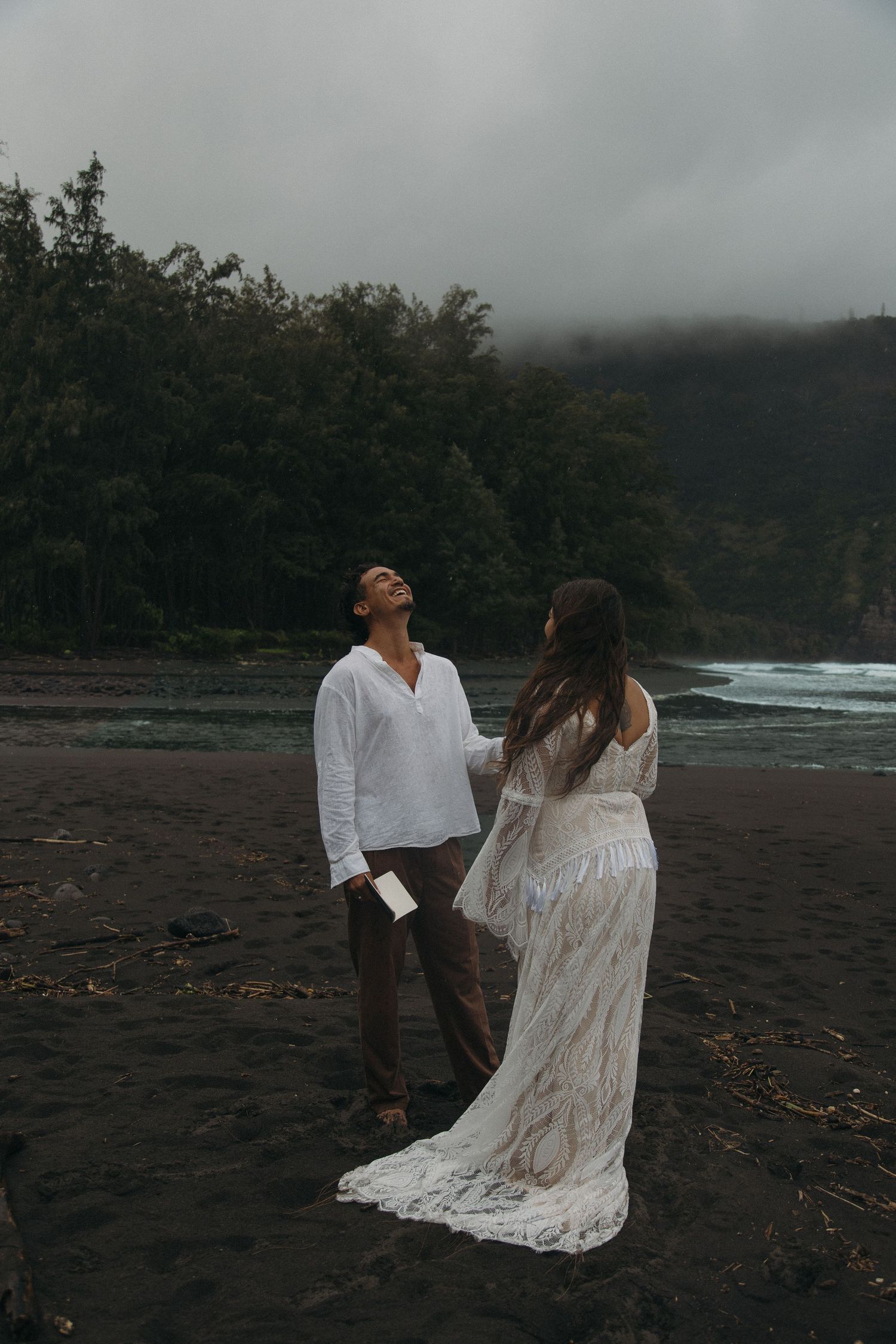 Groom in white shirt laughs joyfully as bride in lace gown approaches on dramatic black sand beach.