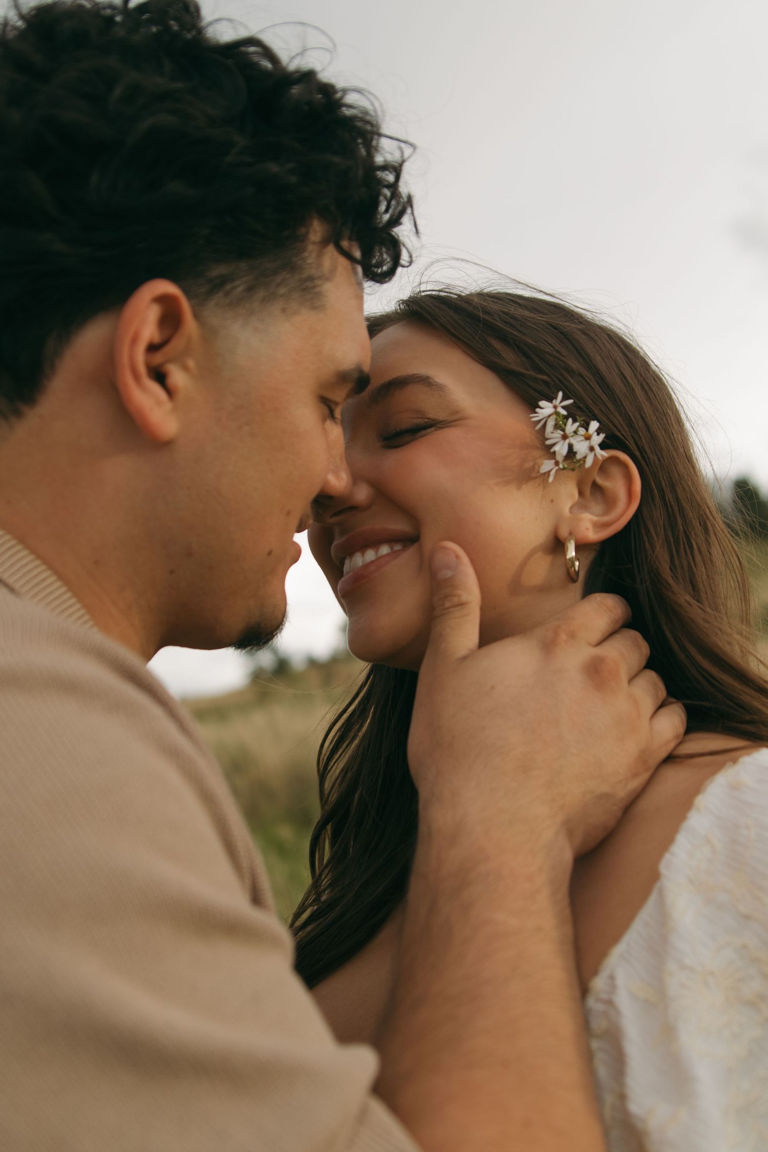 Close-up of smiling couple nearly kissing outdoors, woman wearing floral hair accessories and gold earrings.
