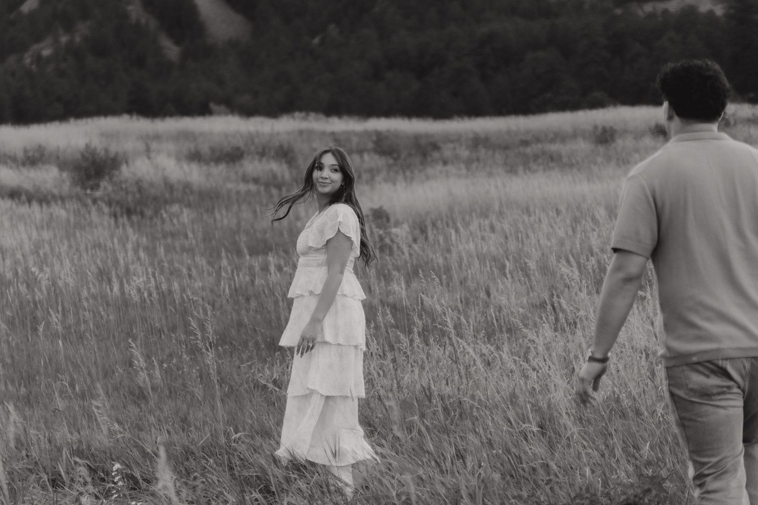 Black and white photo of a woman in a white dress standing in a field, looking back at a man walking toward her.