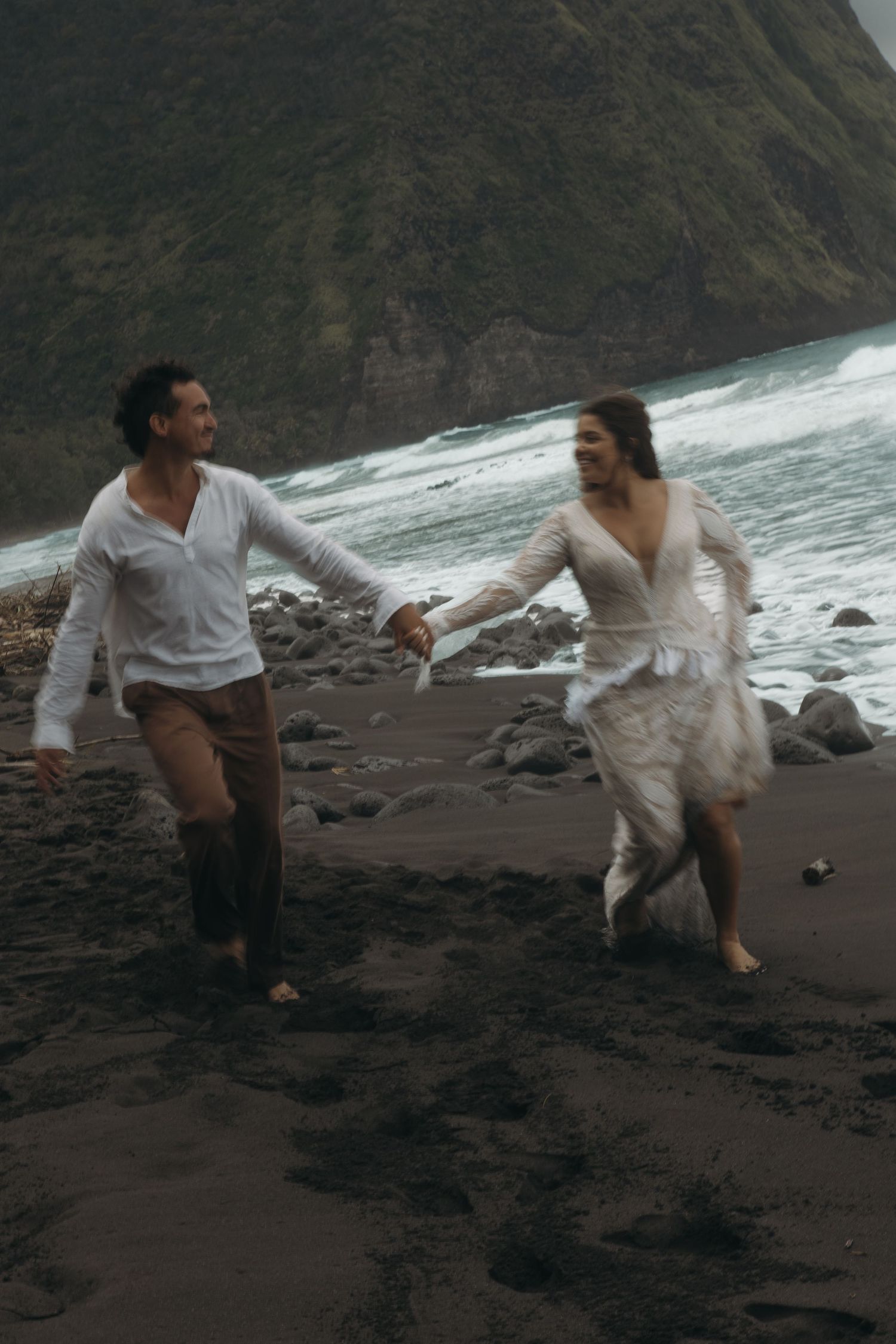 Joyful couple run hand-in-hand along black sand beach with crashing waves and green mountain cliffs.