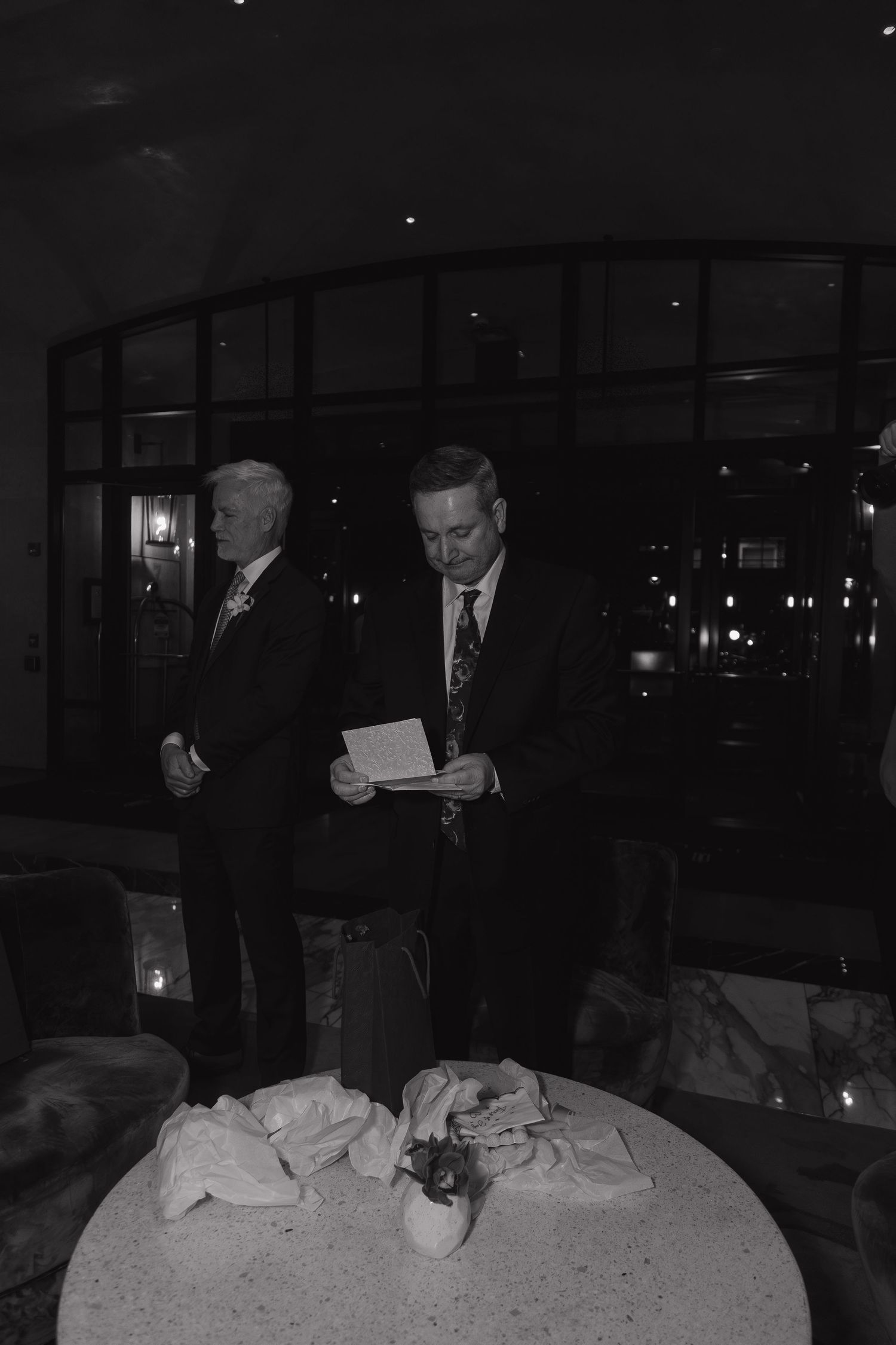 Two men in suits stand near a round table reviewing documents at a formal evening event.