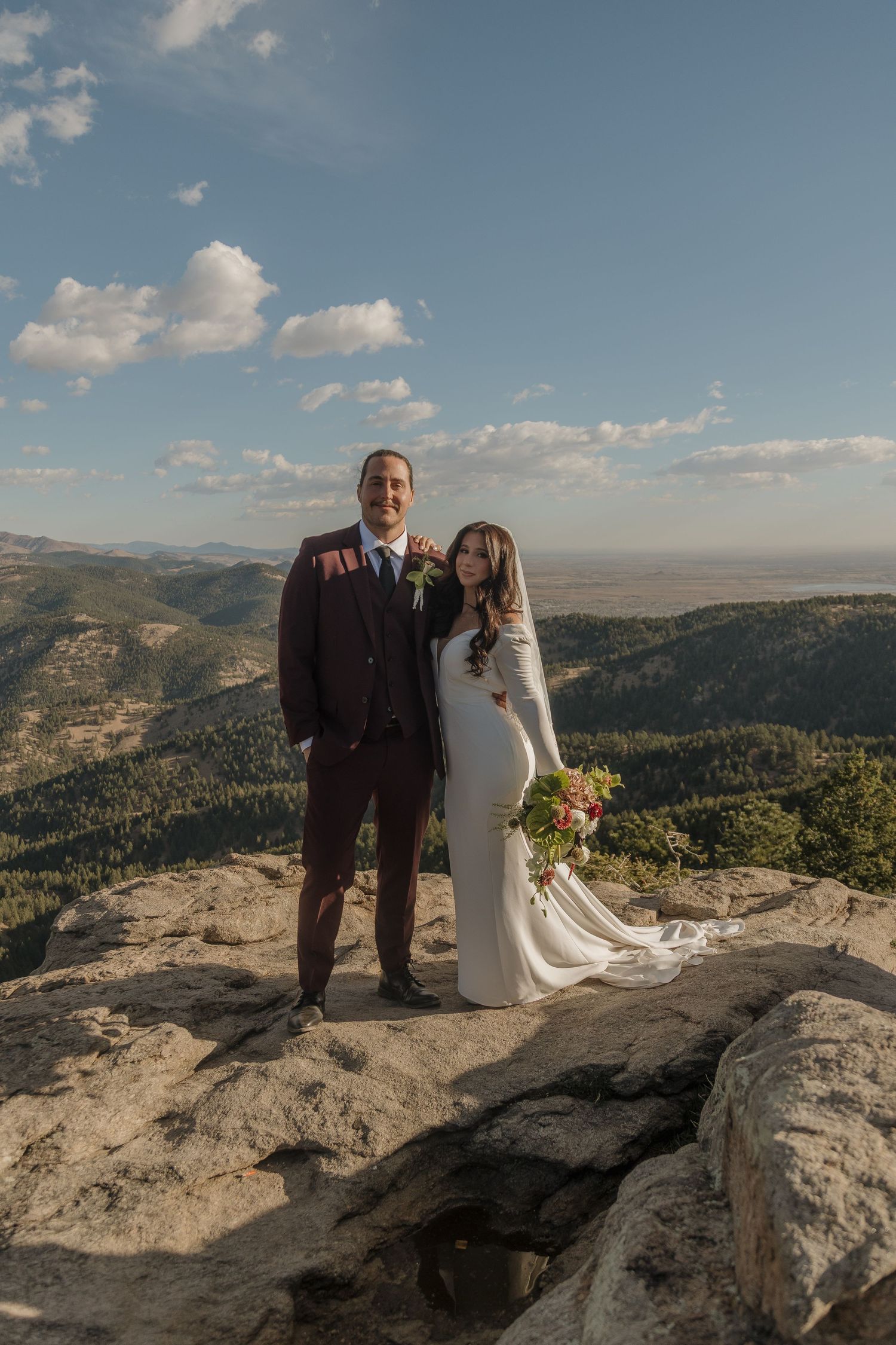 Bride and groom pose together on a mountain overlook, bride holding a colorful bouquet in white gown.