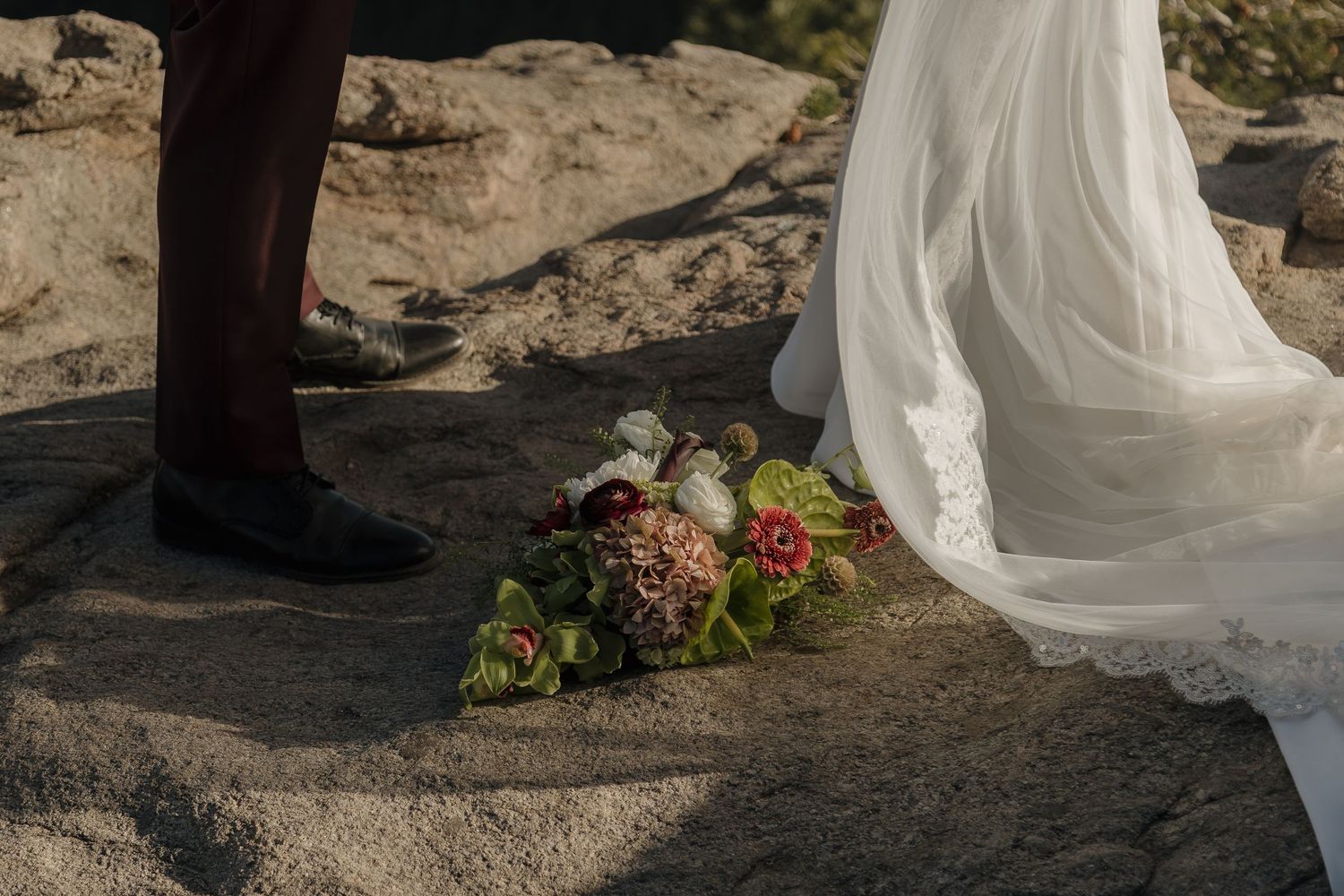 Bridal bouquet of burgundy and green flowers rests on rocky ground beside wedding dress and groom.