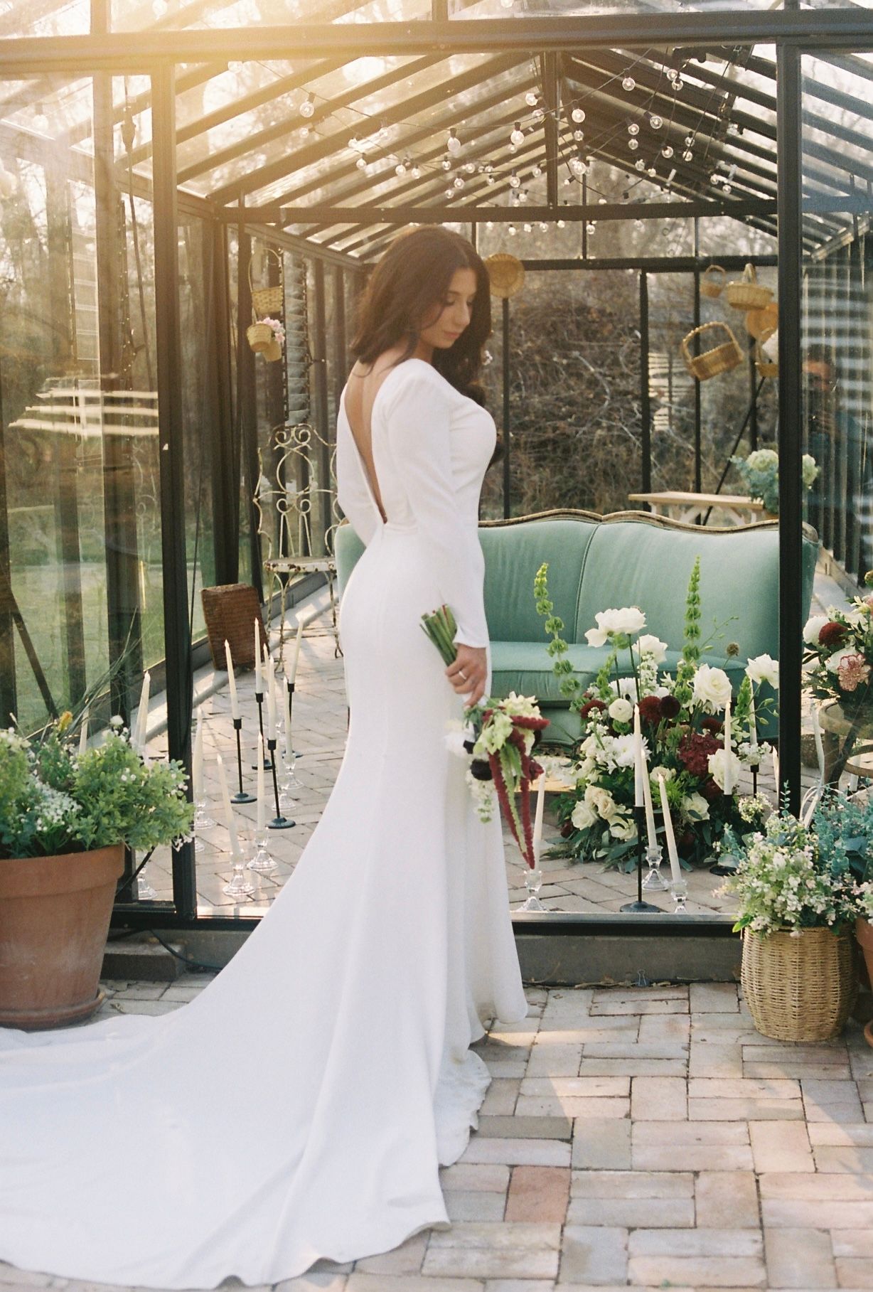 Bride in backless long-sleeve white gown holding bouquet inside sunlit glass greenhouse conservatory.