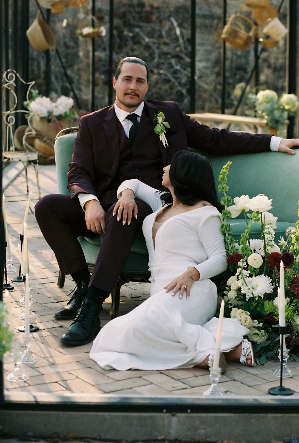 Groom in burgundy suit seated on teal velvet sofa with bride in white gown at decorated greenhouse wedding.