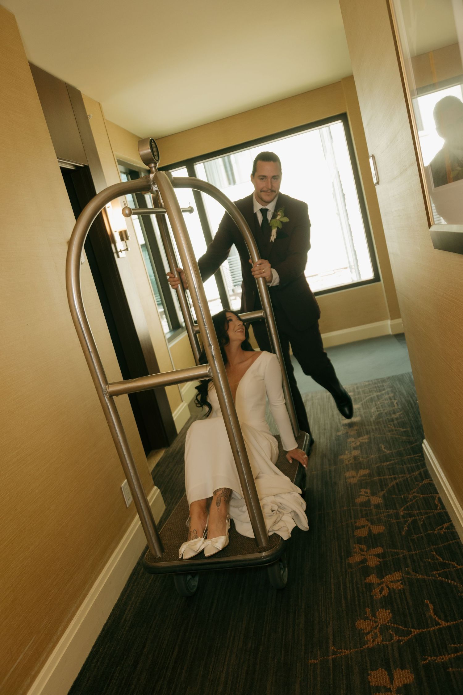 Groom playfully pushes bride on a hotel luggage cart through a carpeted hallway.