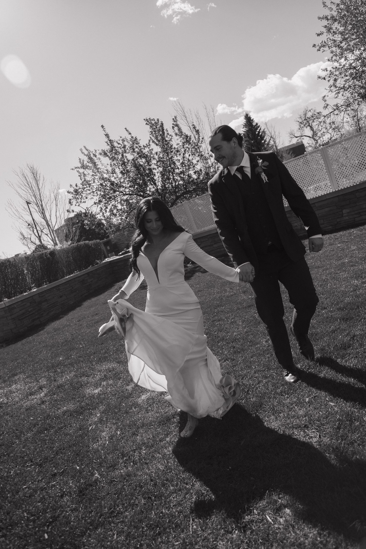 Black and white photo of bride and groom holding hands walking across green lawn on wedding day.