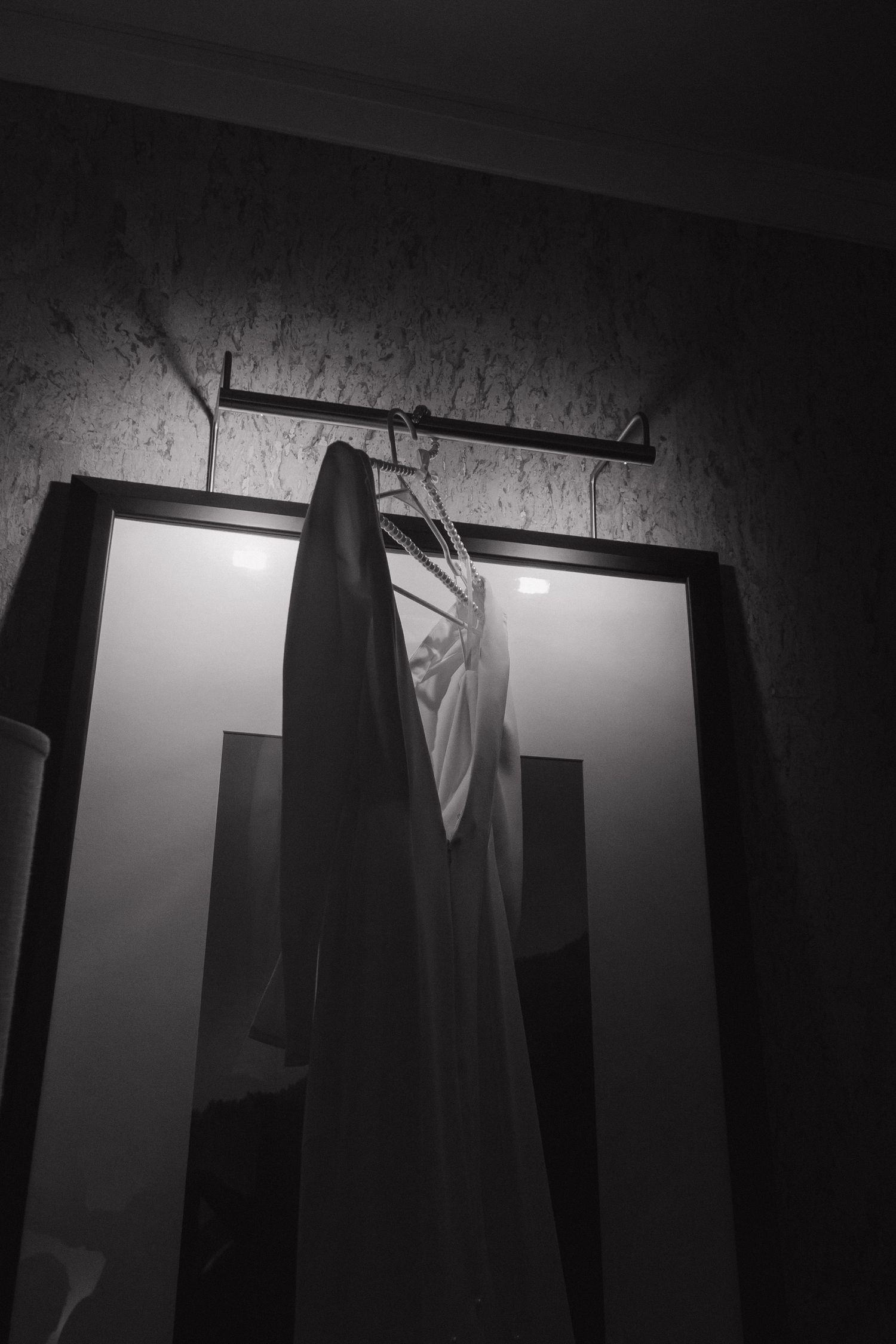 Black and white photo of a white dress hanging on a wall-mounted rack between two mirrors in a dimly lit room.