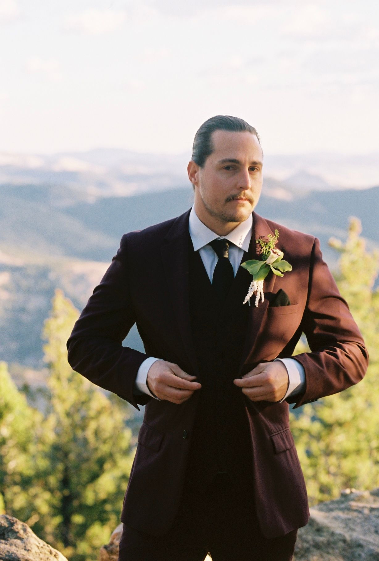 Groom in burgundy three-piece suit adjusts jacket while standing on a rocky overlook with mountain views behind him.