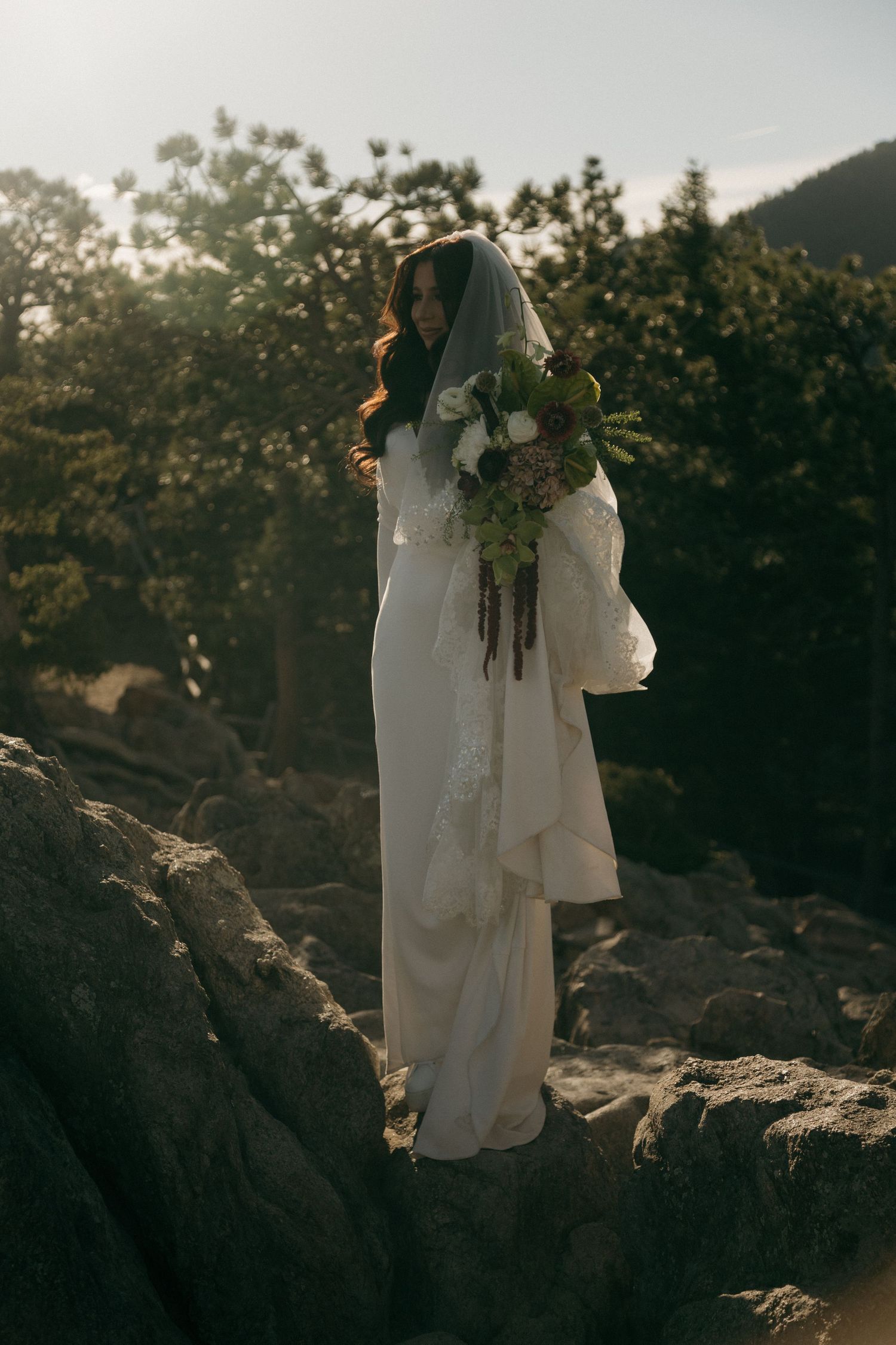 Bride in white gown and veil holds bouquet on rocky mountain terrain with trees in background.